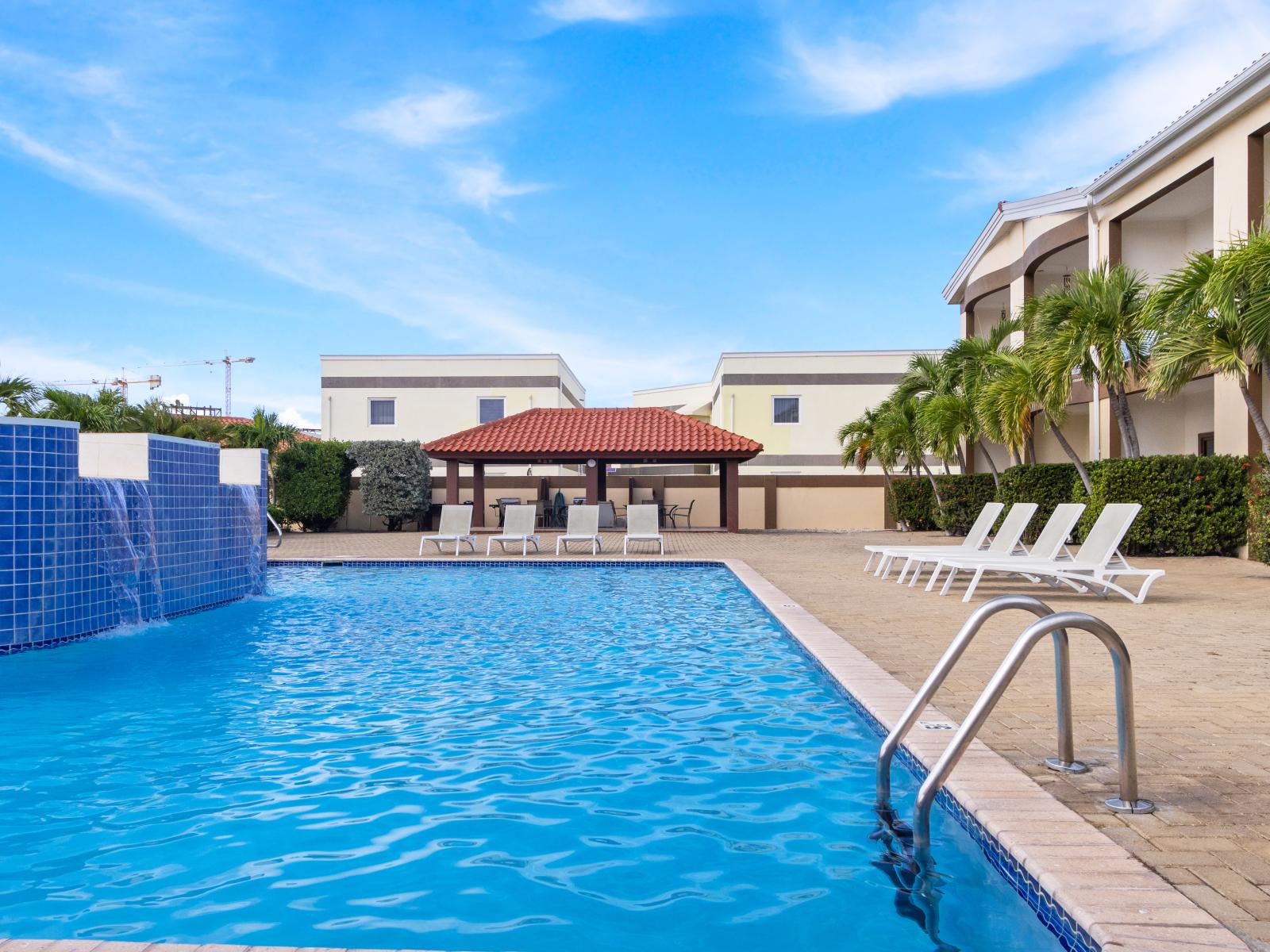 Represent Shared Pool Area of the 2BR Condo in Noord Aruba - Waterfall - Palm trees and tropical plants enhance the vacation feel - Lounge chairs for relaxation