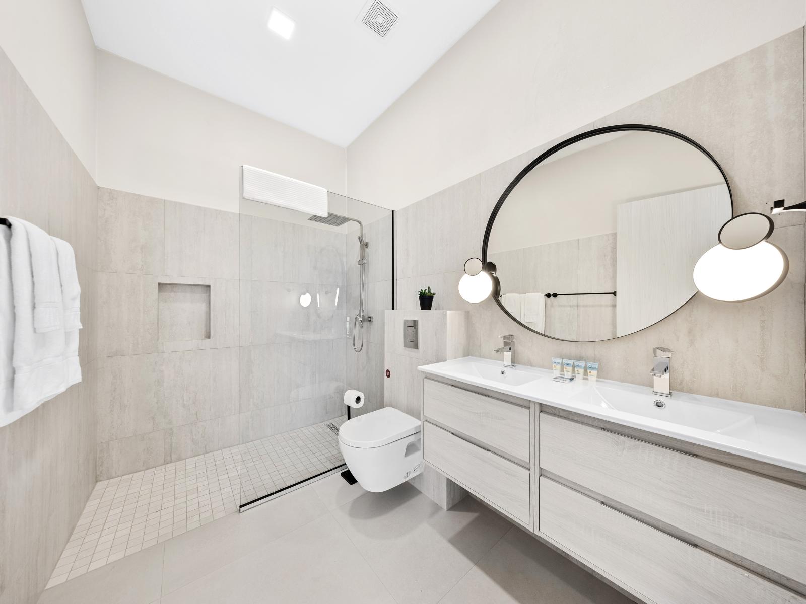 Experience the height of comfort in opulent bathroom of the apartment in Noord Aruba - Features a walk in shower and a luxurious round mirror above the vanity - Enjoy a serene ambiance brought by a sophisticated color palette and upscale lighting