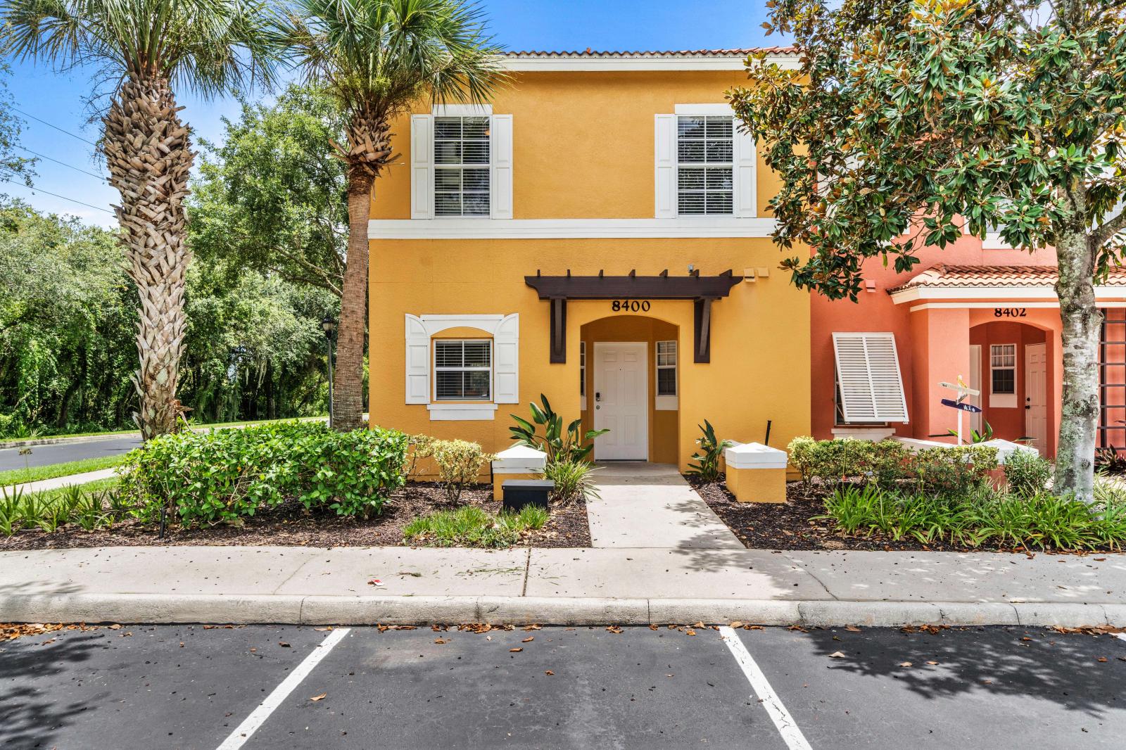 Charming townhouse with a bright yellow facade and white accents, creating a welcoming exterior - Nestled among tall palm trees and lush greenery, providing a tropical ambiance - Convenient parking space located directly in front of the entrance