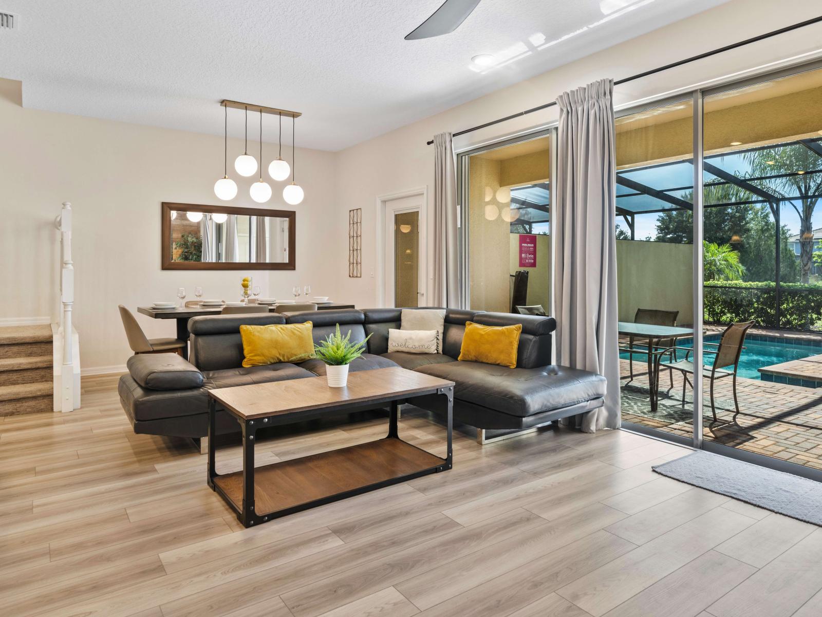 Cozy living area with a chic black sectional and vibrant yellow pillows - Modern dining space featuring an elegant light fixture and large mirror - Smart TV and inviting patio with a sparkling pool just beyond sliding glass doors