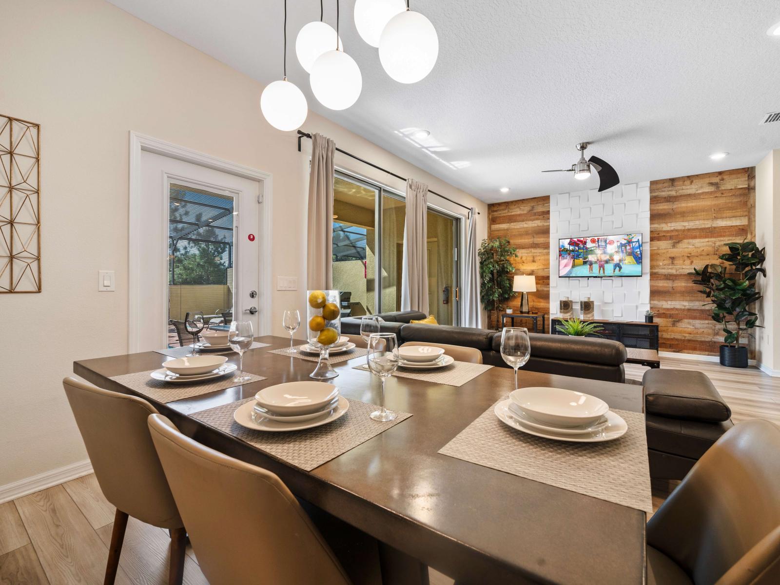 Open-plan dining area with modern furnishings, flooded with natural light from large window - An elegantly set dining table and a cozy living area with a wall-mounted TV - Sliding glass doors, creating a seamless indoor-outdoor living experience