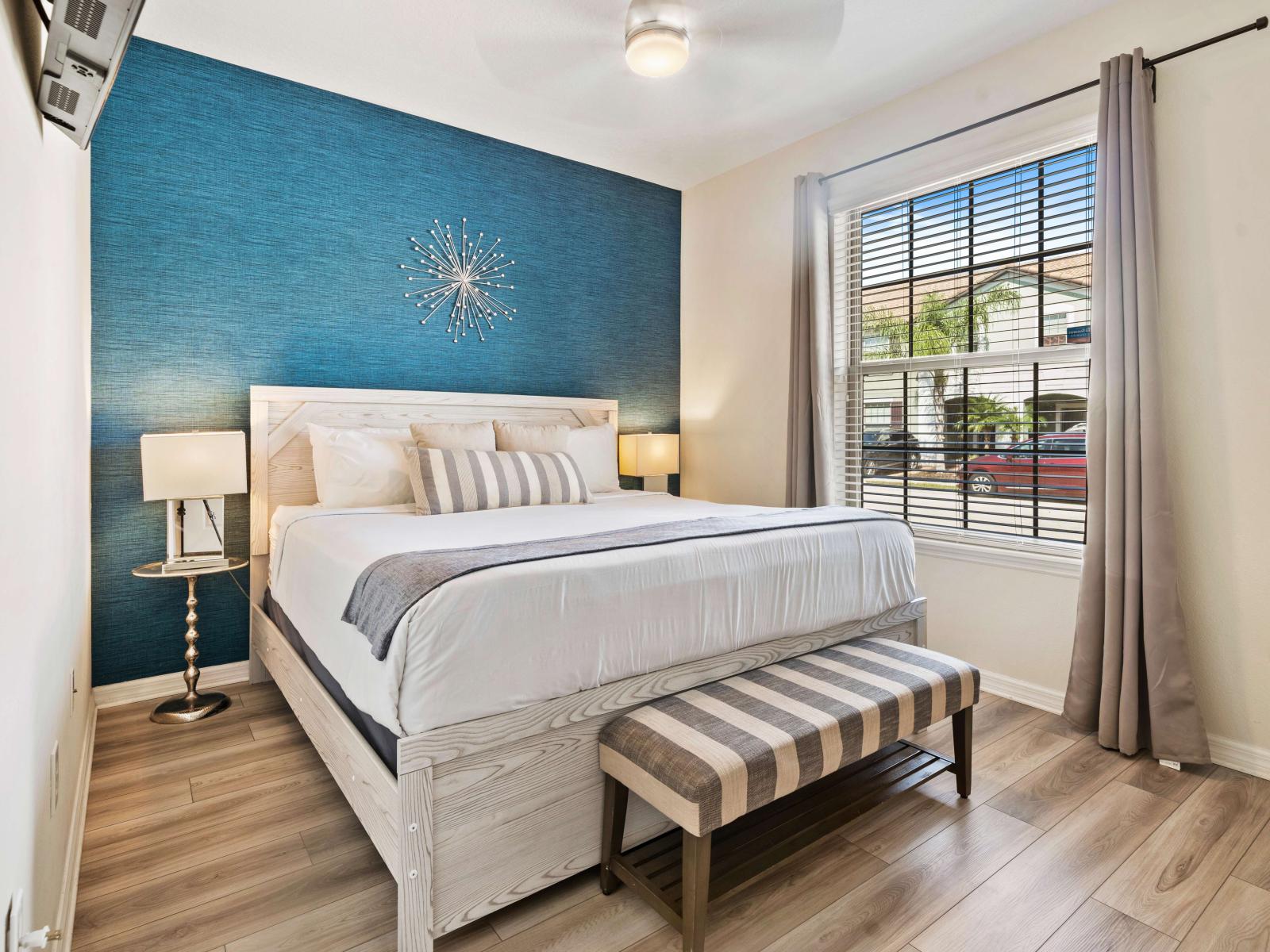 A King-size bed with elegant bedding and decorative pillows - The room is highlighted by a blue accent wall, and a stylish bench at the foot of the bed - A large window offering plenty of natural light