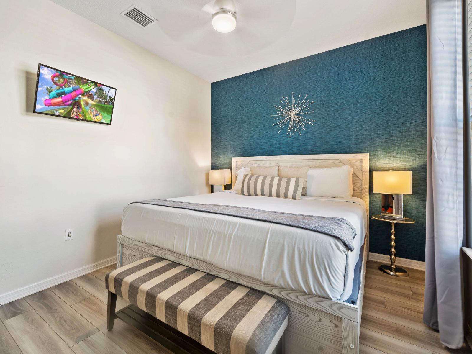 A king-size bed with a stylish wooden frame and a striped bench at its foot - The room features a striking teal accent wall with modern starburst decor - A smart TV and elegant bedside lamps on round tables provide entertainment and a cozy ambiance