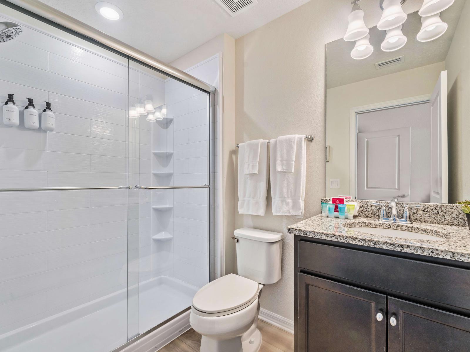 Walk-in shower with sleek glass doors, equipped with built-in shelves and a dispenser - The elegant granite countertop on the single sink vanity offers space - The bathroom is well-appointed with fresh white towels and essential toiletries