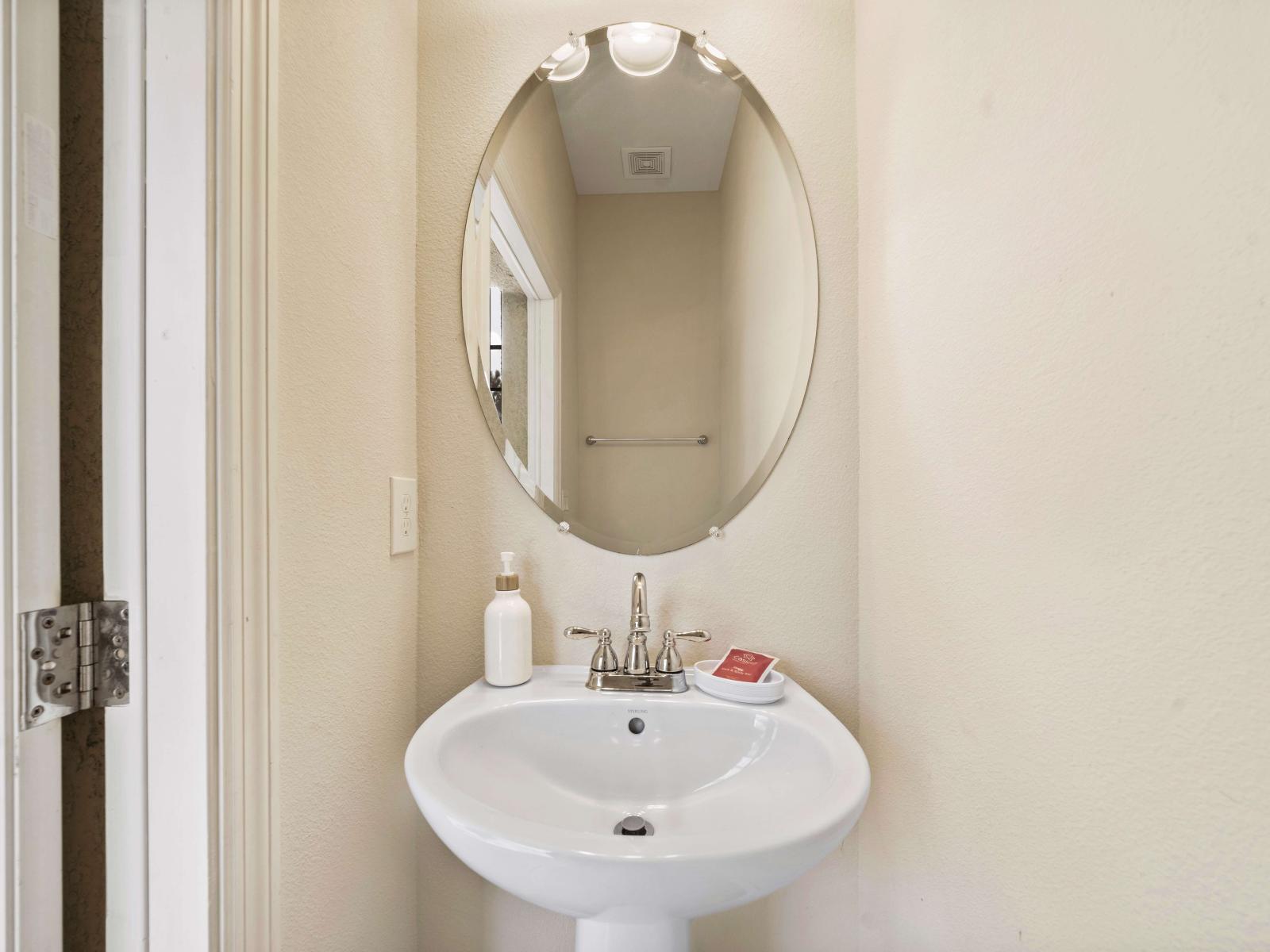 The half bathroom features with a sleek pedestal sink and an elegant oval mirror - Toiletries are neatly arranged on the sink, providing convenience for guests - The compact space is well-lit, ensuring for quick freshening up