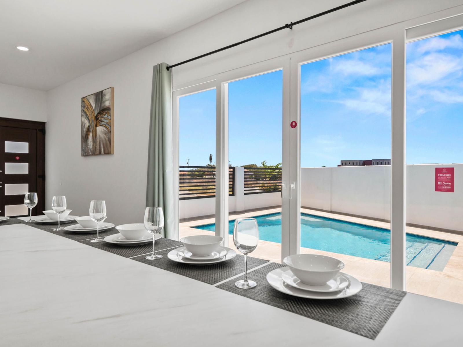 - A stunning view of a private pool from the dining area, for an indoor-outdoor feel - Elegant table setting ready for a meal, with white dishware and crystal-clear glasses - Bright, open space complemented by modern decor and a serene outdoor oasis