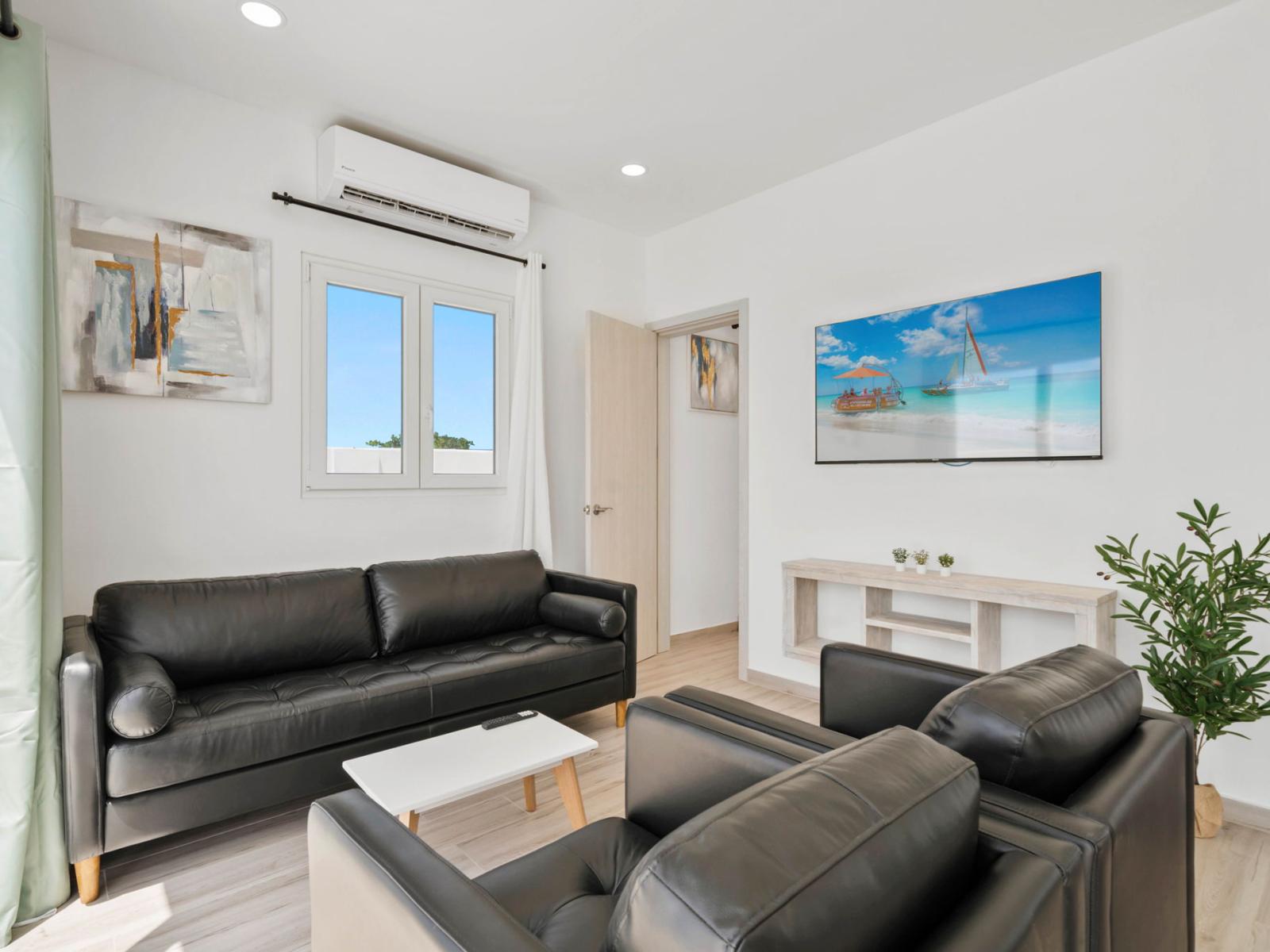 - Cozy living room with sleek black leather seating and a minimalist design - Wall-mounted TV displays a vibrant beach scene, adding a touch of relaxation to the space - Natural light and simple decor create a bright, airy atmosphere for lounging