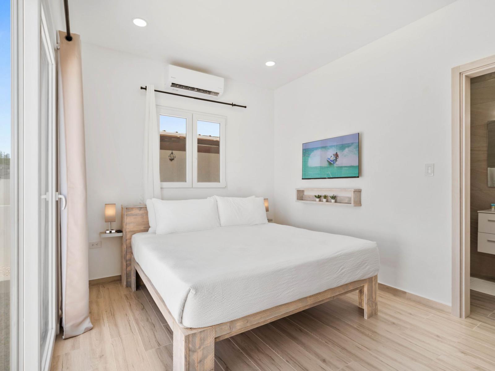 - Bright and airy bedroom featuring a comfortable bed with crisp white linens - Minimalist decor with a wall-mounted TV and soft lighting create a relaxing ambiance - Sliding glass doors open to the outdoor space, blending indoor and outdoor living