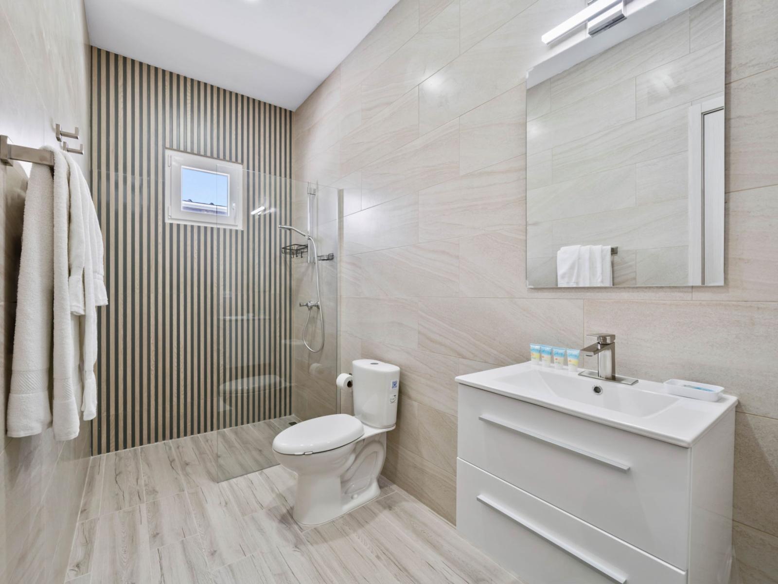 - Modern bathroom with sleek, light-toned tiles and a walk-in shower with a glass partition - Minimalist vanity with storage space, complemented by a large mirror - Elegant striped accent wall adds a stylish touch to the clean, contemporary design