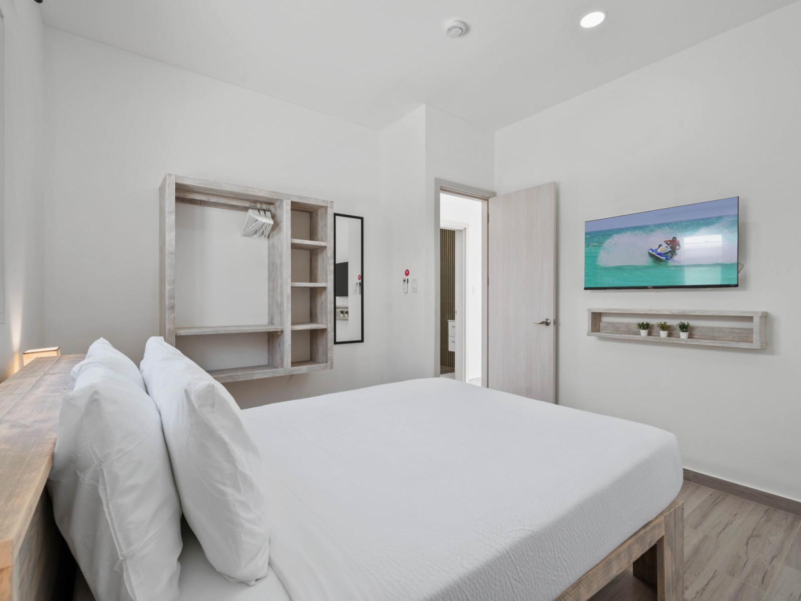 - Cozy bedroom with a queen-size bed and crisp white linens for a restful atmosphere - Open shelving and a wall-mounted TV add functionality and entertainment to the design - Bright natural light enhances the room’s minimalist, serene design