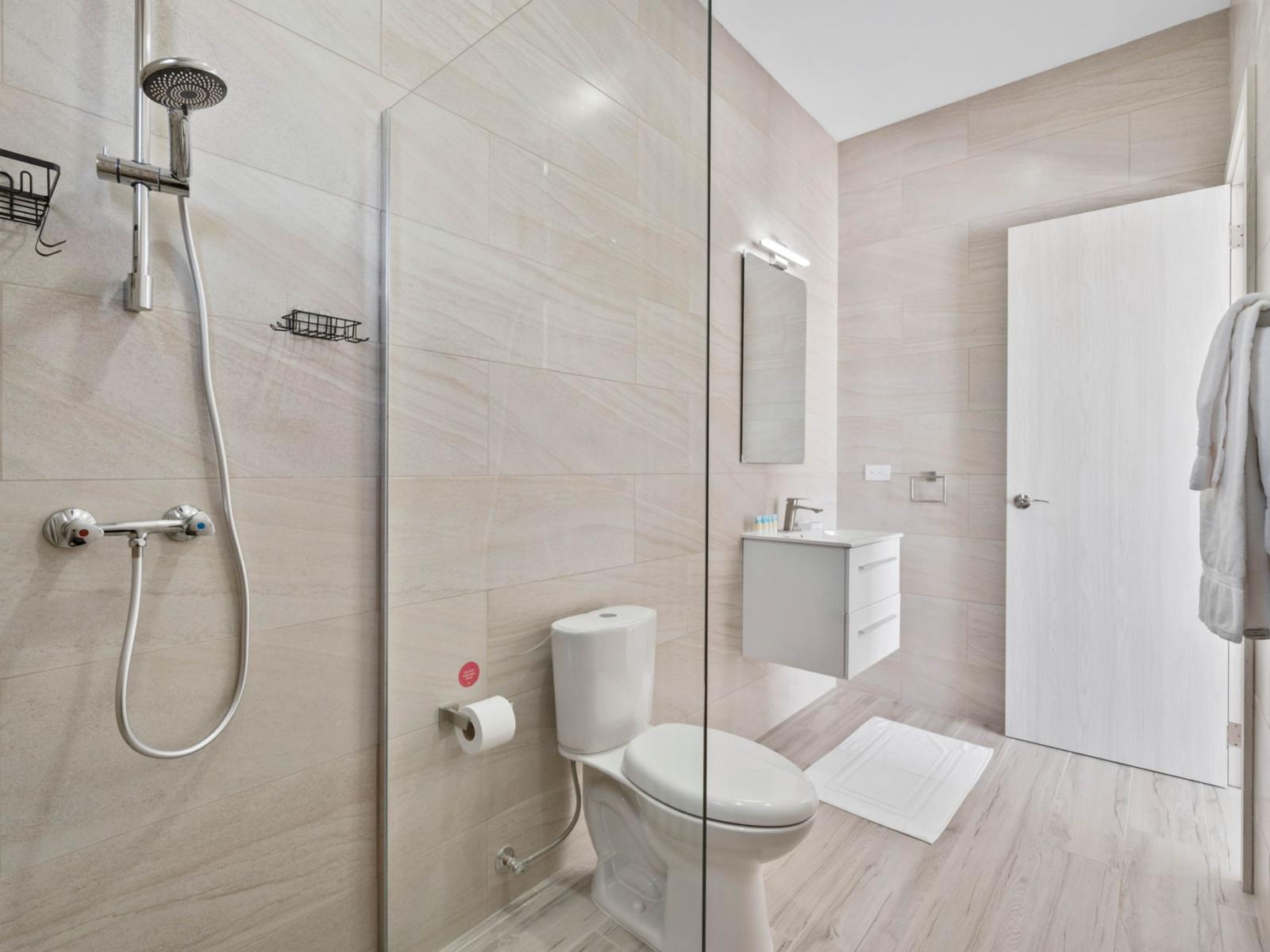 - Modern bathroom with sleek neutral tiles and a glass-enclosed walk-in shower - Floating vanity and bright lighting create a clean, spacious feel   - Simple, minimalist design with functional storage and fresh white towels for added comfort