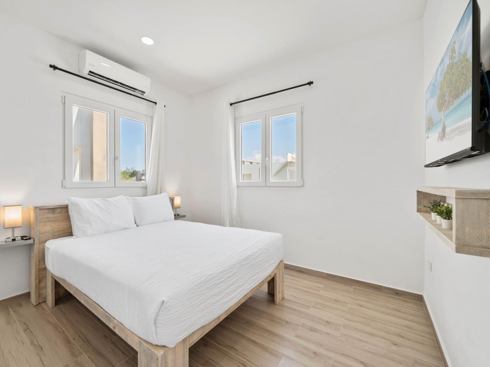 - Bright bedroom with a queen-size bed, featuring crisp white linens for a clean look - Dual windows allow natural light to flood the space, enhancing the room’s fresh ambiance - Wall-mounted TV and small shelf with plants add a cozy, modern touch