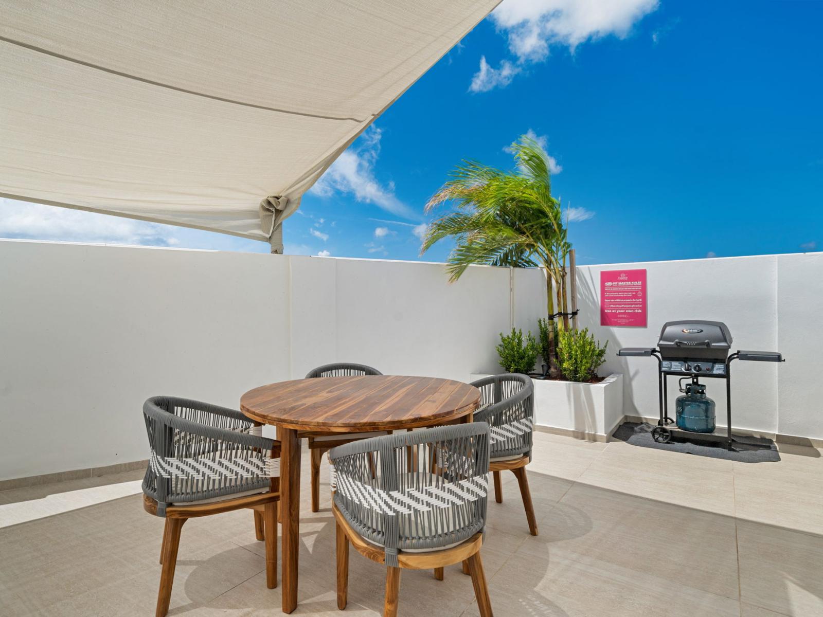 - Outdoor patio area with a wooden round table and comfortable seating, perfect for dining al fresco - Gas grill nearby for easy outdoor cooking, ideal for barbecues and casual meals - Tropical plants enhance the relaxing and inviting atmosphere