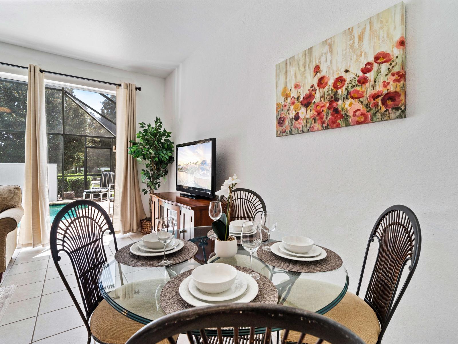 Charming dining area with a glass table set for four, perfect for intimate meals - Large windows provide plenty of natural light and a view of the private pool - Decorative floral artwork and a TV for entertainment enhance the dining experience