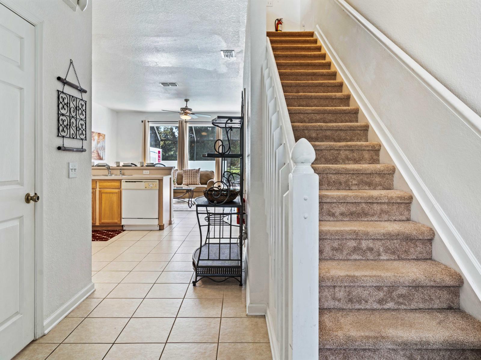 Open living area with a view of the kitchen and cozy seating - Carpeted staircase leading to the upper level, adding a touch of warmth and comfort - Functional kitchen space with essential appliances and a clear sightline to the living room