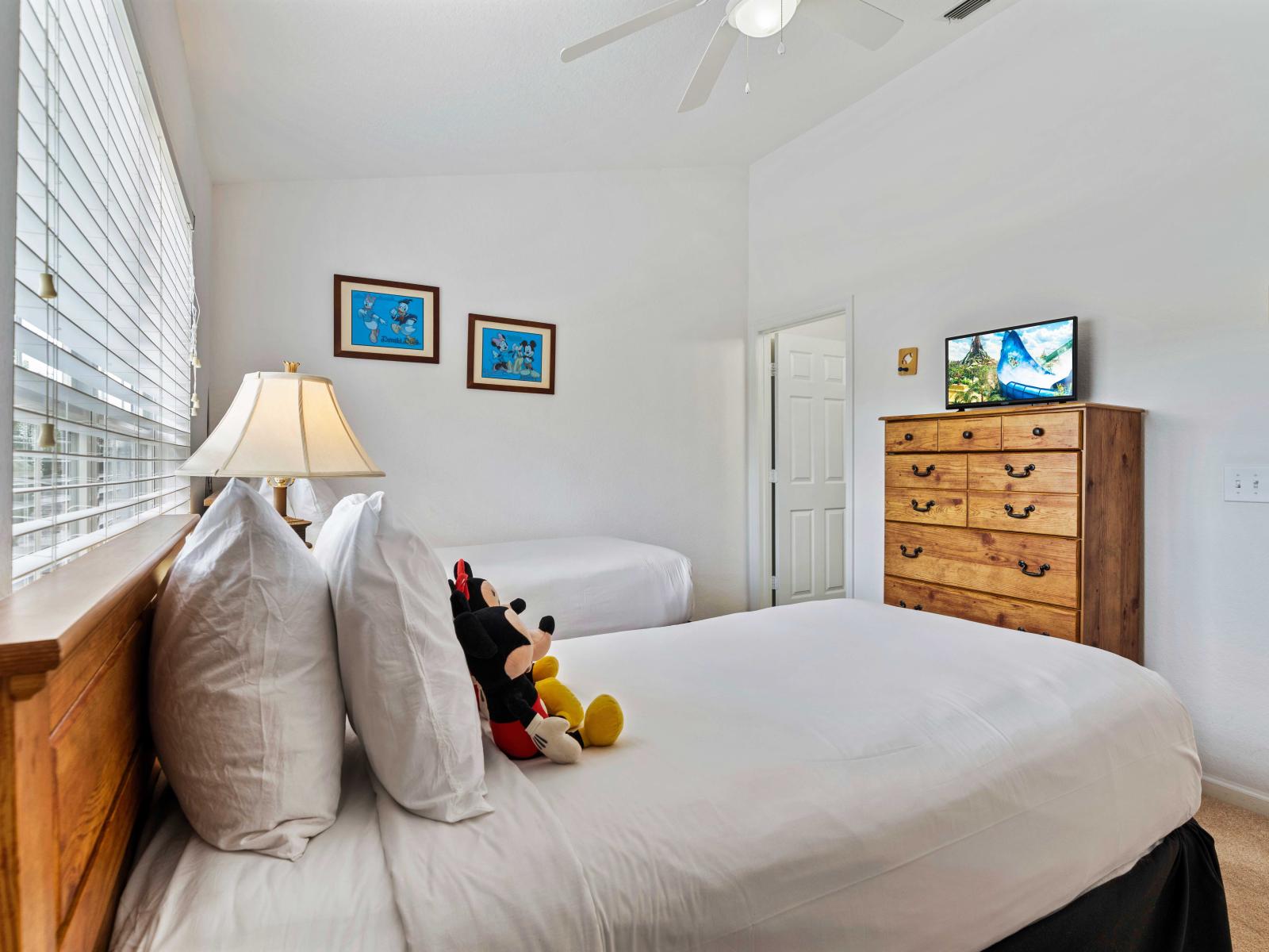 Bright and inviting bedroom with two single beds, perfect for kids - Decorated with Disney-themed artwork and plush toys, creating a playful and cheerful environment - Features a TV and storage space, ensuring comfort and entertainment for all