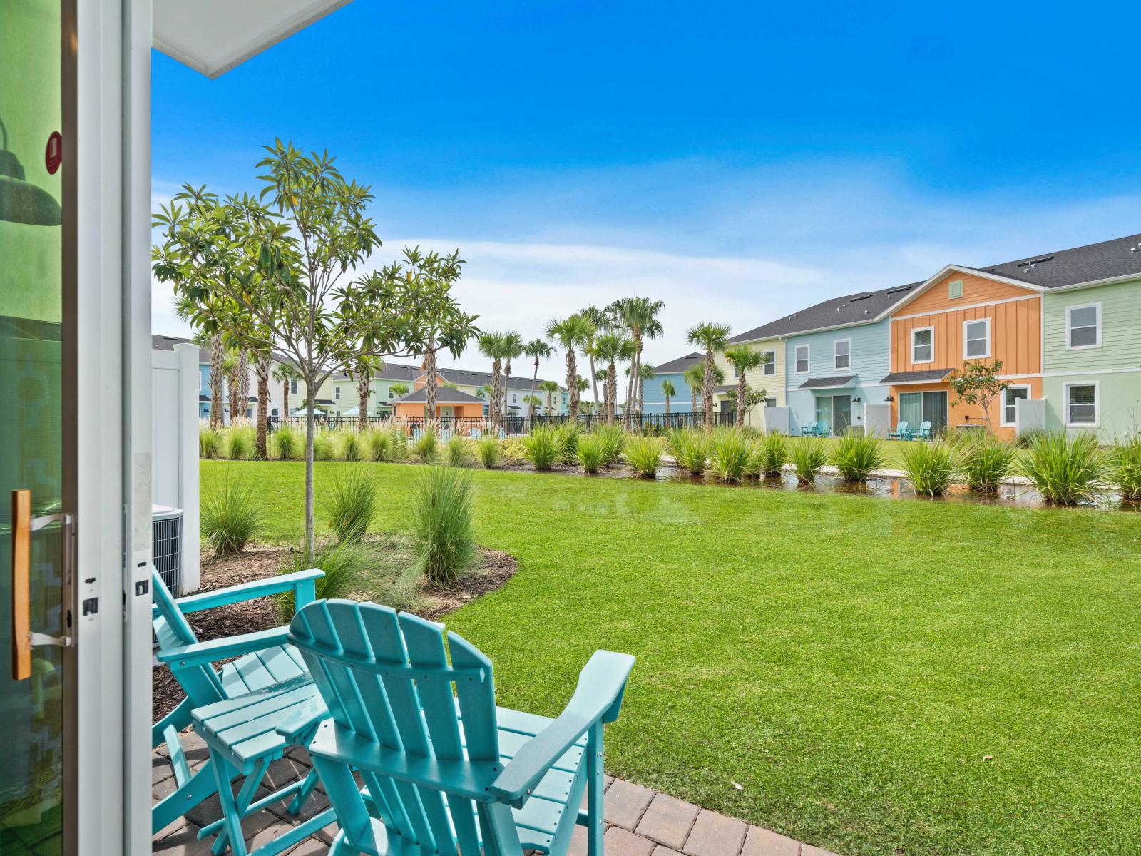 Cozy patio with turquoise Adirondack chairs, ideal for morning coffee or evening relaxation - Lush green lawn and scenic area with palm trees and ornamental grasses, creating a tranquil retreat - Colorful townhouses, adding vibrant charm