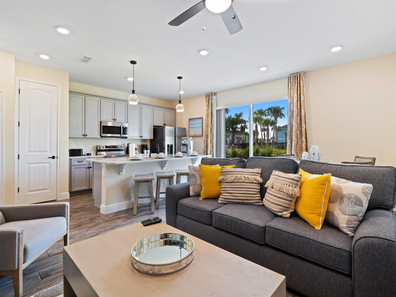 Stylish living area featuring a modern kitchen with stainless steel appliances and a breakfast bar with stools - Comfortable gray sofa adorned with yellow and patterned pillows - Large sliding glass door revealing a scenic view of sunny outdoors