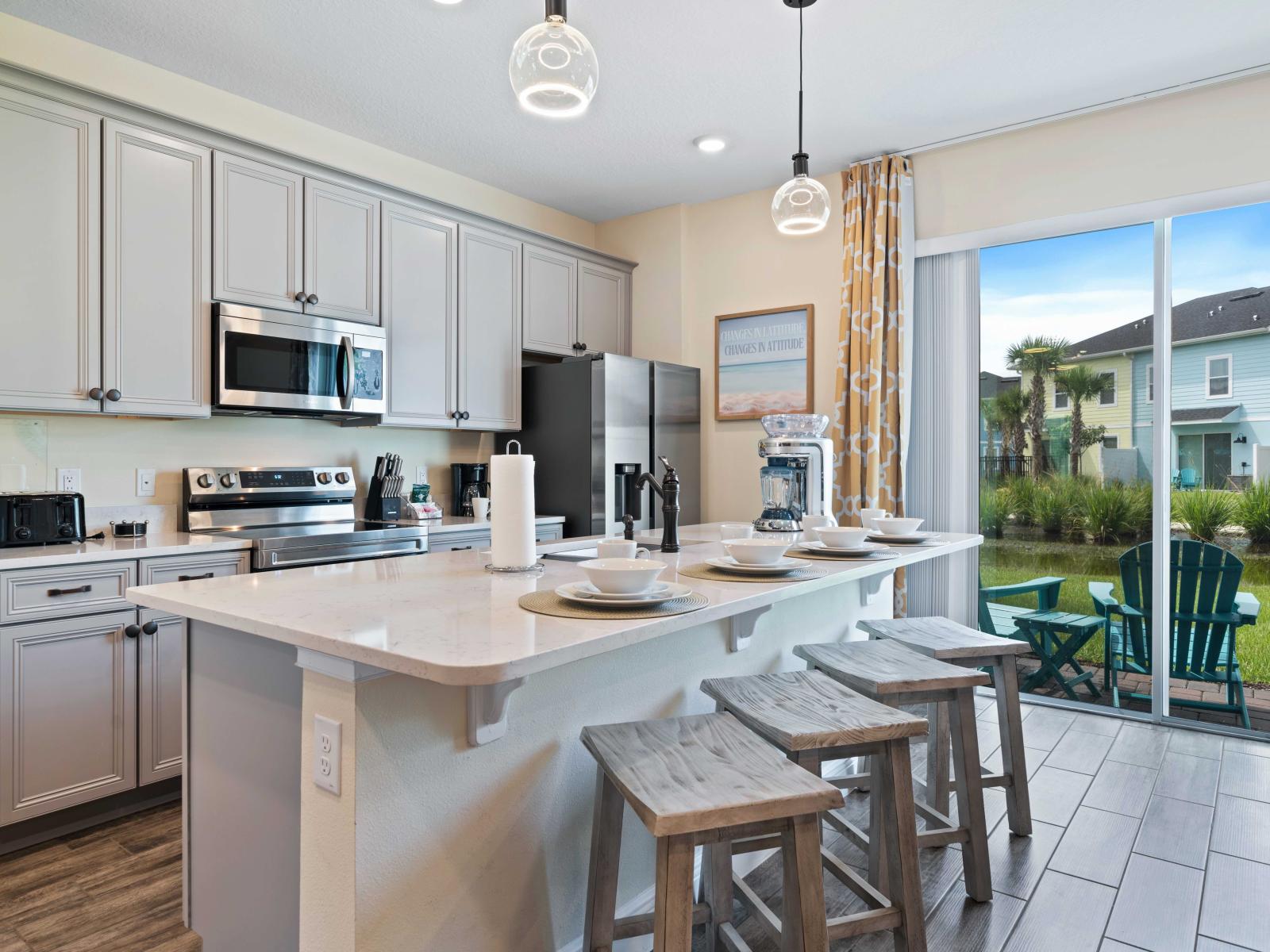 Modern, fully equipped kitchen featuring stainless steel appliances and cabinetry - Large island with a white marble countertop, seating for four, and stylish pendant lighting - Sliding glass door provides plenty of natural light with a scenic view