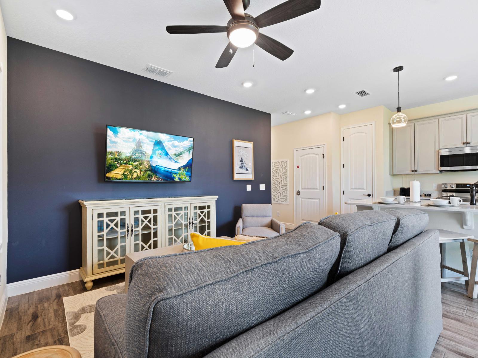 Bright and welcoming living room with a plush gray sofa and a stylish armchair - Navy accent wall features a mounted flat-screen TV above a vintage-style cabinet - Open floor plan flows into a contemporary kitchen and a cozy breakfast bar