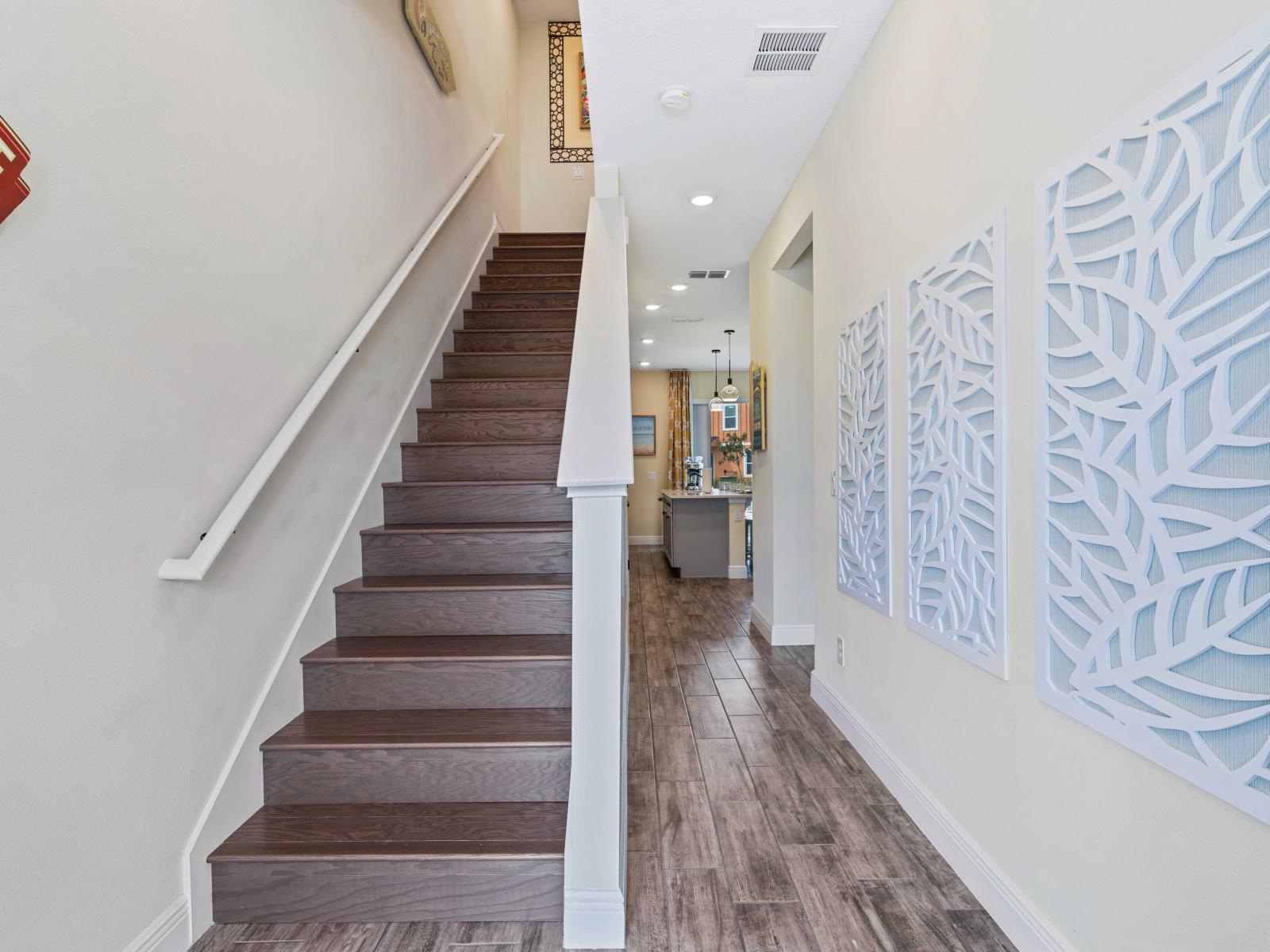 Inviting entry hallway featuring elegant wooden stairs and modern decor - White wall art with intricate leaf designs adds a stylish touch - Leads into an open kitchen and living area, creating a seamless flow throughout the home