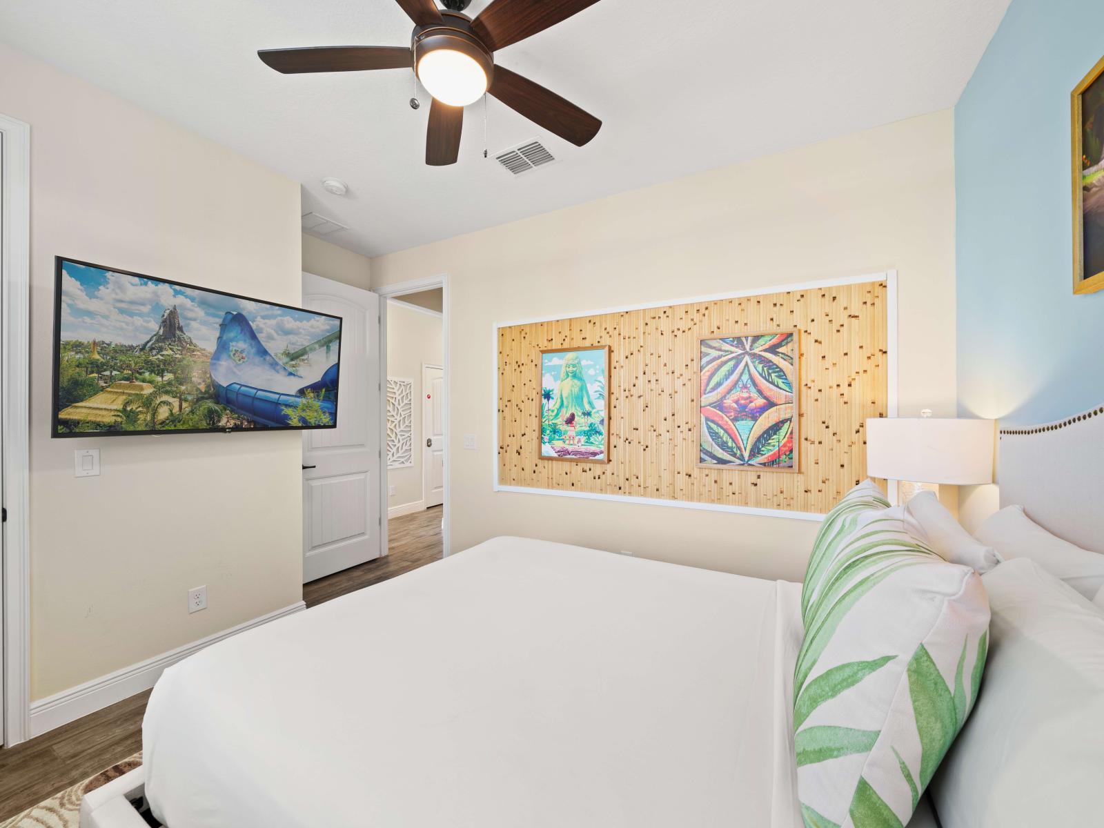 Bright bedroom featuring a queen-sized bed with leaf-patterned pillows and white bedding - Large windows with light blue curtains allow natural light and offer an outdoor view - Wall-mounted TV for your entertainment, adding a vibrant touch