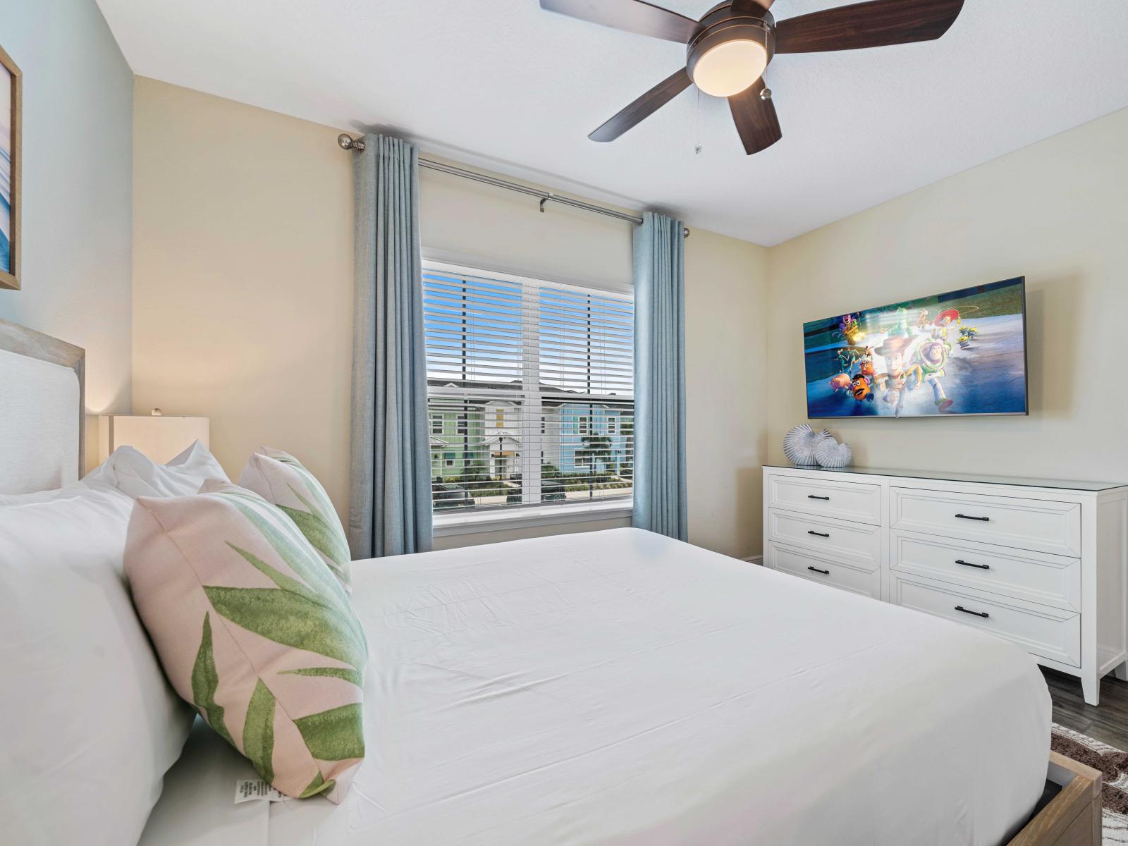 Inviting bedroom with a queen-sized bed, adorned with leaf-patterned pillows and white bedding - Large window with light blue curtains allows natural light and offers a pleasant view - Features a wall-mounted TV complemented by a white dresser