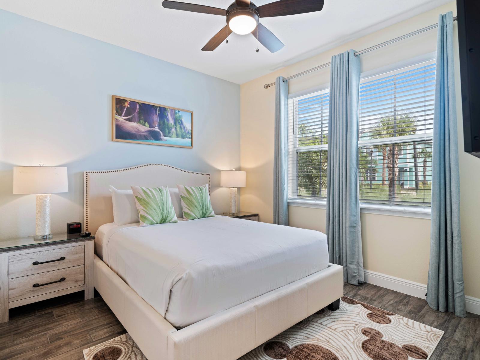 Cozy bedroom with a queen-sized bed, leaf-patterned pillows, and white bedding - Natural light from large windows with light blue curtains brightens the room - Features matching bedside tables, modern lamps, and a serene wall painting