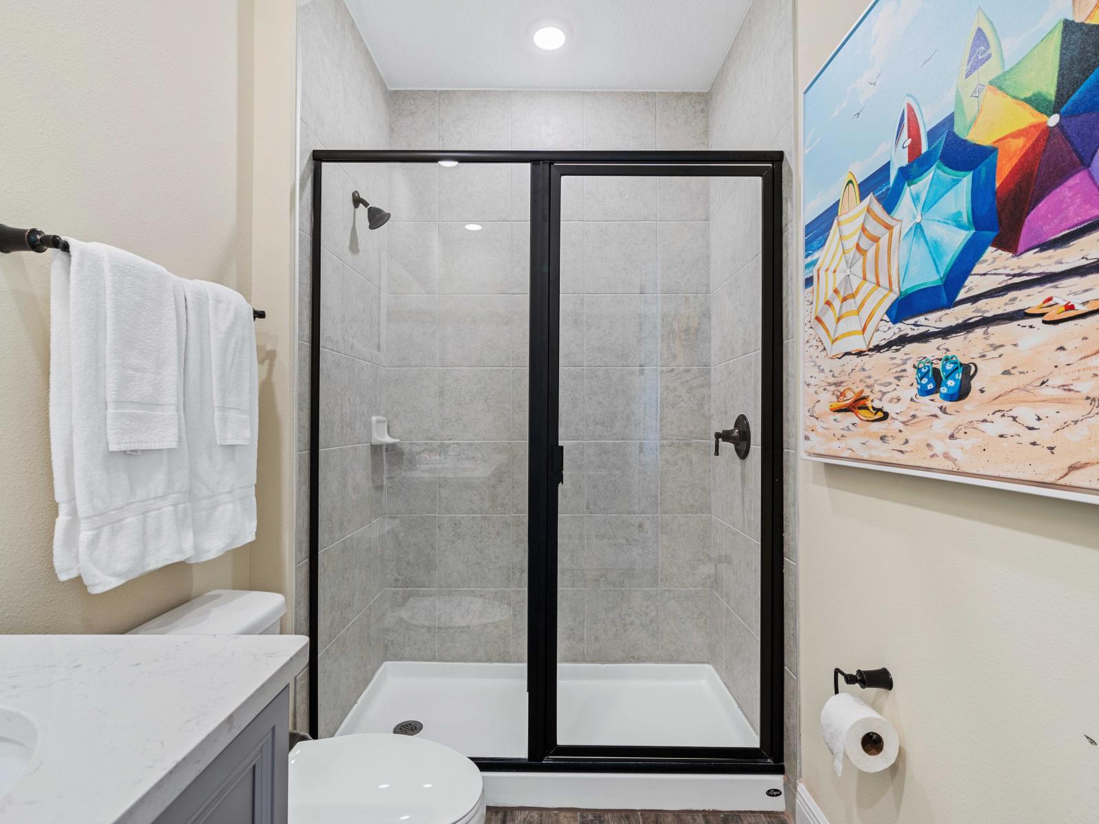 Elegant bathroom with a walk-in shower enclosed by black-framed glass doors - Features a white marble countertop, black fixtures, and neatly hung white towels - Bright space enhanced by colorful beach-themed wall art, adding a vibrant touch