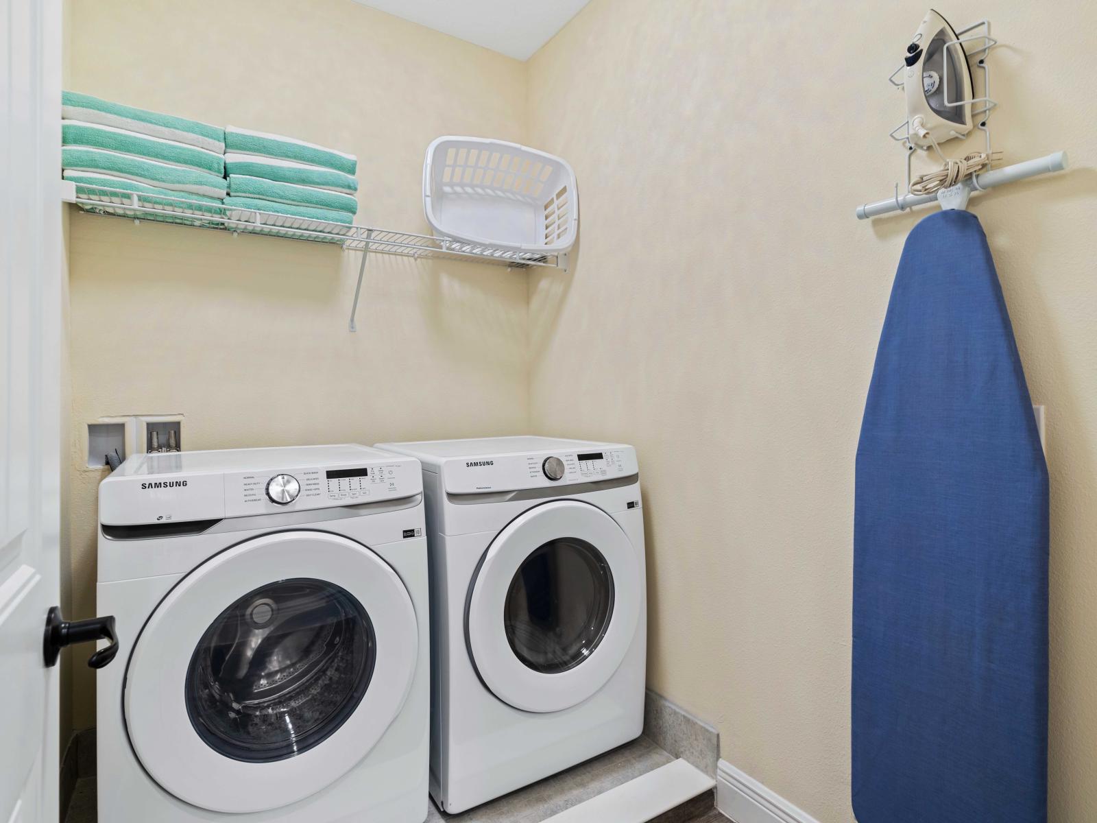 Laundry room equipped with a washer and dryer for convenient in-home laundry care - Wire shelf above the machines holds neatly folded towels and a laundry basket - Ironing board and iron mounted on the wall for easy access and storage