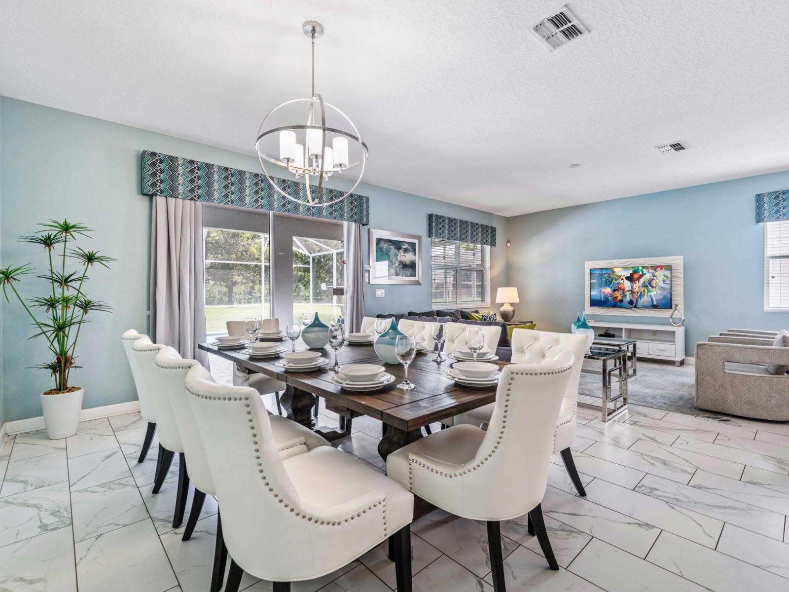 - Spacious dining area with a large wooden table set for eight persons - Modern chandelier and decorative curtains enhancing the room's elegance - Bright and airy with a view of the outdoor space through sliding glass door