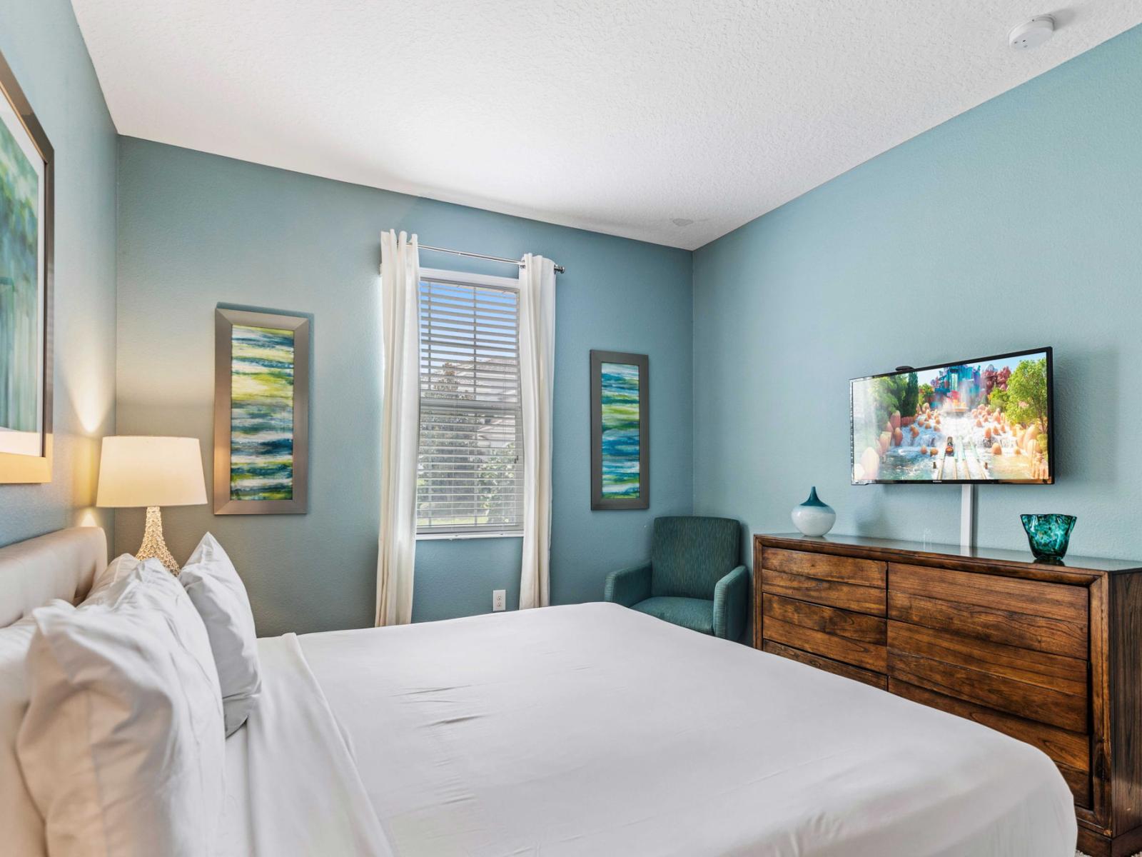 - Bedroom is centered around a plush king bed dressed in crisp white linens - Aesthetic is accentuated by a soft blue accent wall and vibrant, colorful wall art - Additional features include a stylish wooden dresser, smart TV and comfortable armchair