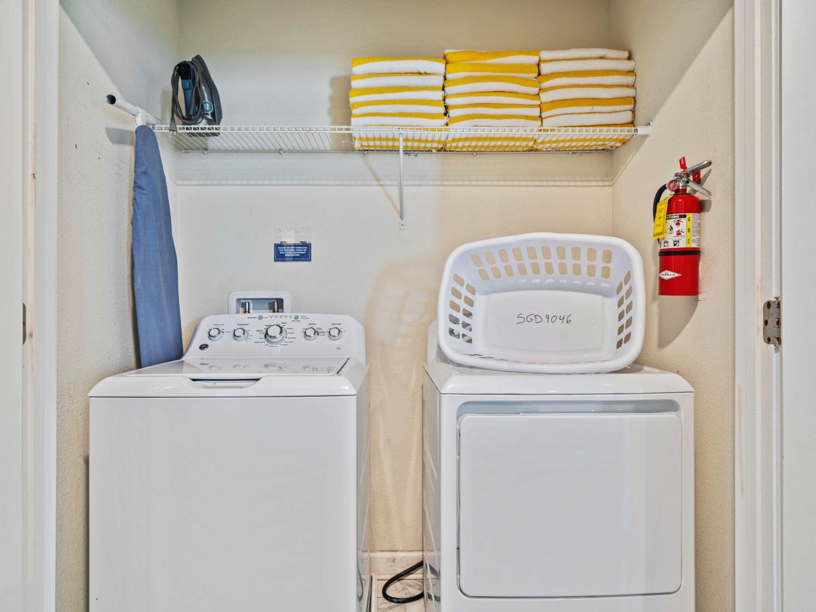 - Washer and dryer set with a clean and organized layout - Shelving above the machines stocked with neatly folded yellow and white towels - Essential laundry items like an iron, ironing board, and laundry basket within reach