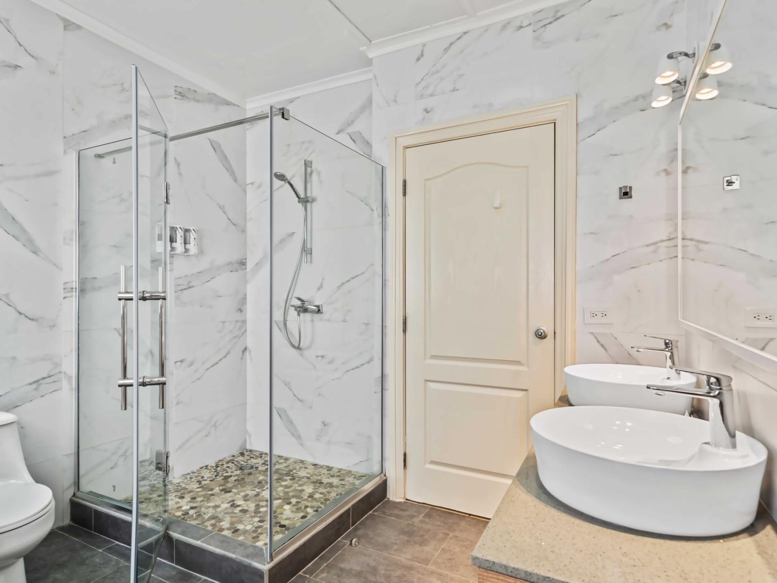 Modern walk-in shower with glass doors and a rainfall showerhead - The shower area has a pebble-tile floor, adding a touch of natural texture - The sleek, white toilet and dual sinks with chrome fixtures enhance the bathroom's modern aesthetic