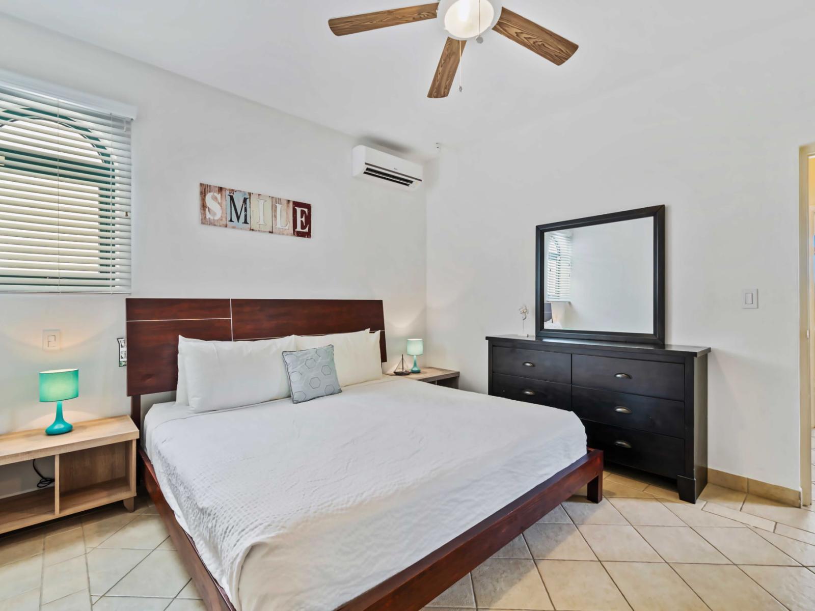 Spacious king-size bed with a sleek wooden headboard - The room features a large dresser with a mirror, providing storage and a place to get ready - Air conditioning unit ensure comfort, while the cheerful "SMILE" wall decor adds a positive touch