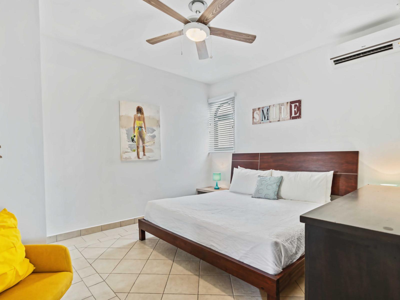 Bedroom features a king-size bed with a modern wooden headboard - Bright yellow accent chair and beach-themed wall art add personality and a touch of color - Natural light fills the room through the window, enhancing the airy atmosphere