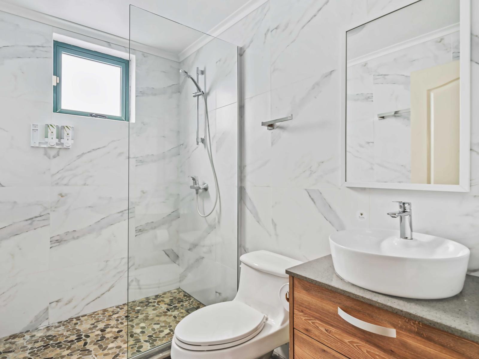 A modern walk-in shower with a floor designed with pebble tiles, adding a stylish touch - Bathroom includes a sleek white bowl sink atop a wooden vanity with storage space - Elegant marble-patterned tiles cover the walls, providing a peaceful feel