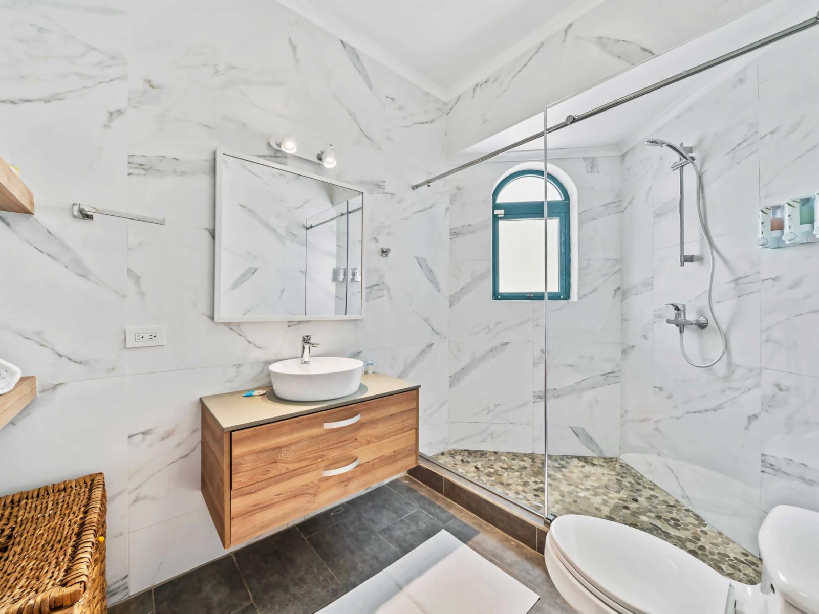 The shower area, accented with pebble-tile flooring, adding a natural and modern touch - An arched window allows natural light to flood the space, adding to its airy atmosphere - Elegant marble-patterned tiles cover the walls, enhancing space