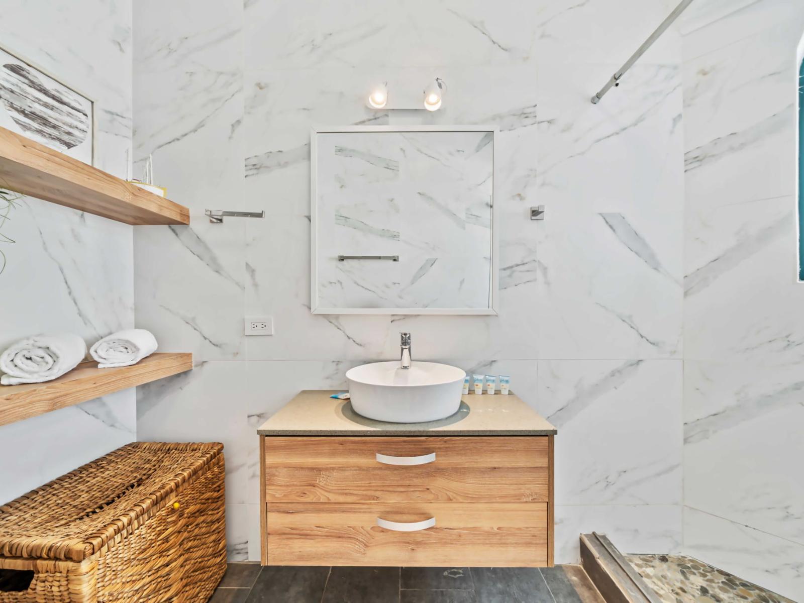 The sleek, bowl sink sits atop a floating wooden vanity with drawers for storage - Open wooden shelves provide storage, while a wicker basket adds a rustic element - Mirror, complemented by wall-mounted lighting, enhances the brightness of the room