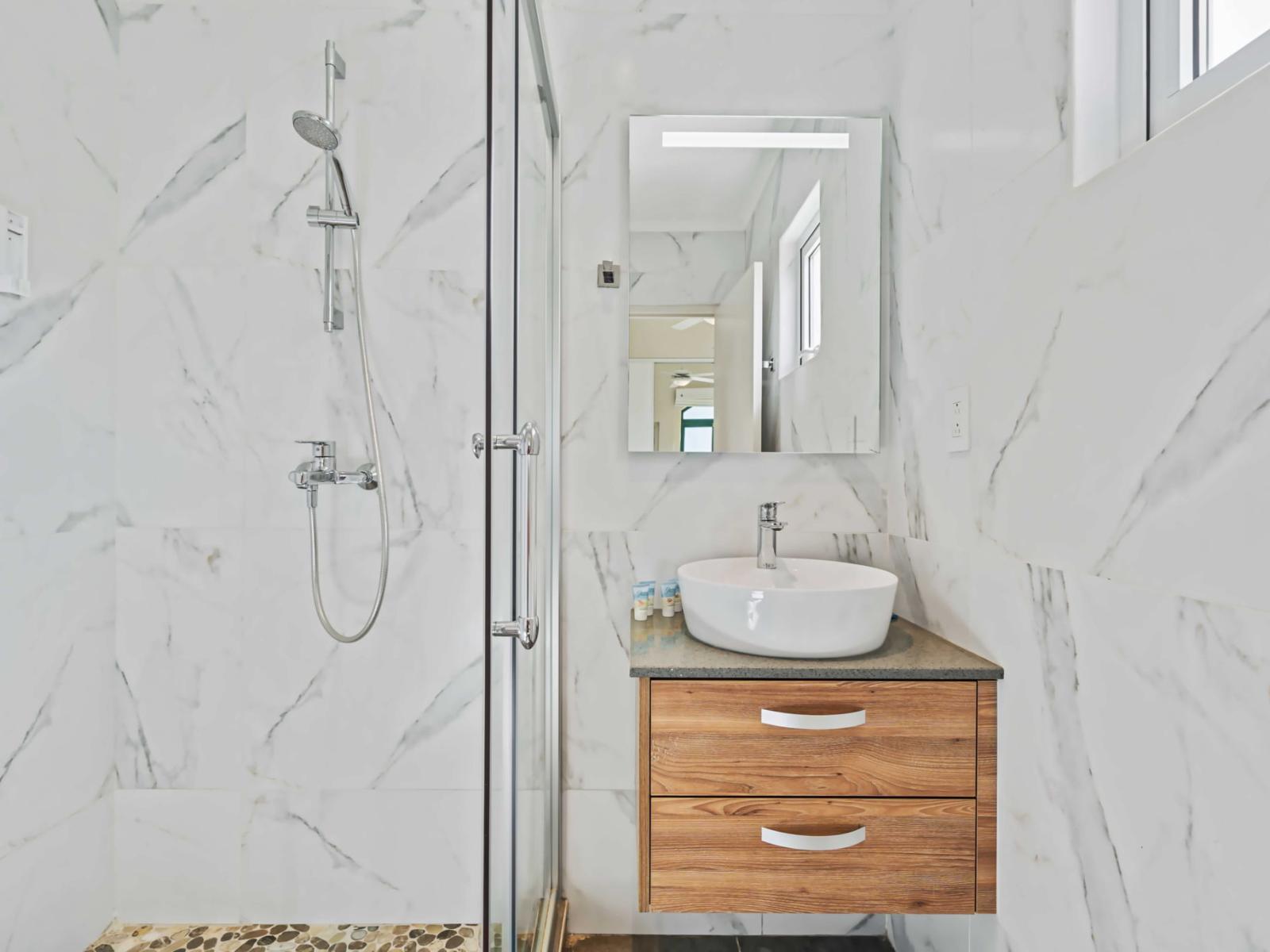 The bathroom features a sleek walk-in shower with glass doors and a modern rainfall showerhead - Marble-patterned tiles adorn the walls, giving the space a cohesive look - A white bowl sink is mounted on a wooden vanity, providing storage