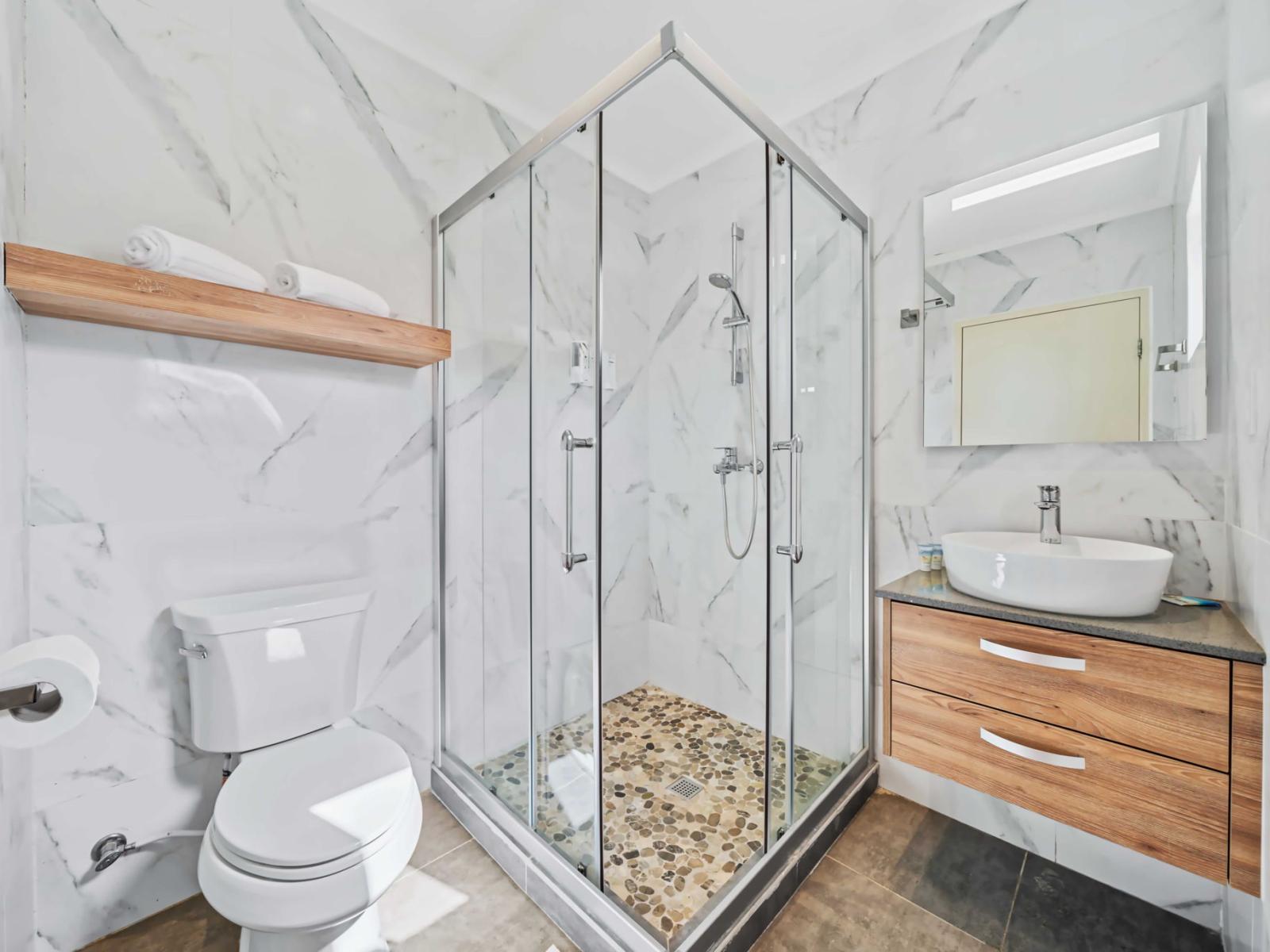 The bathroom includes a walk-in shower with glass doors and a contemporary rainfall showerhead - An open wooden shelf above the toilet provides extra space for towels and decorative items - A sleek white bowl sink sits on a floating wooden vanity