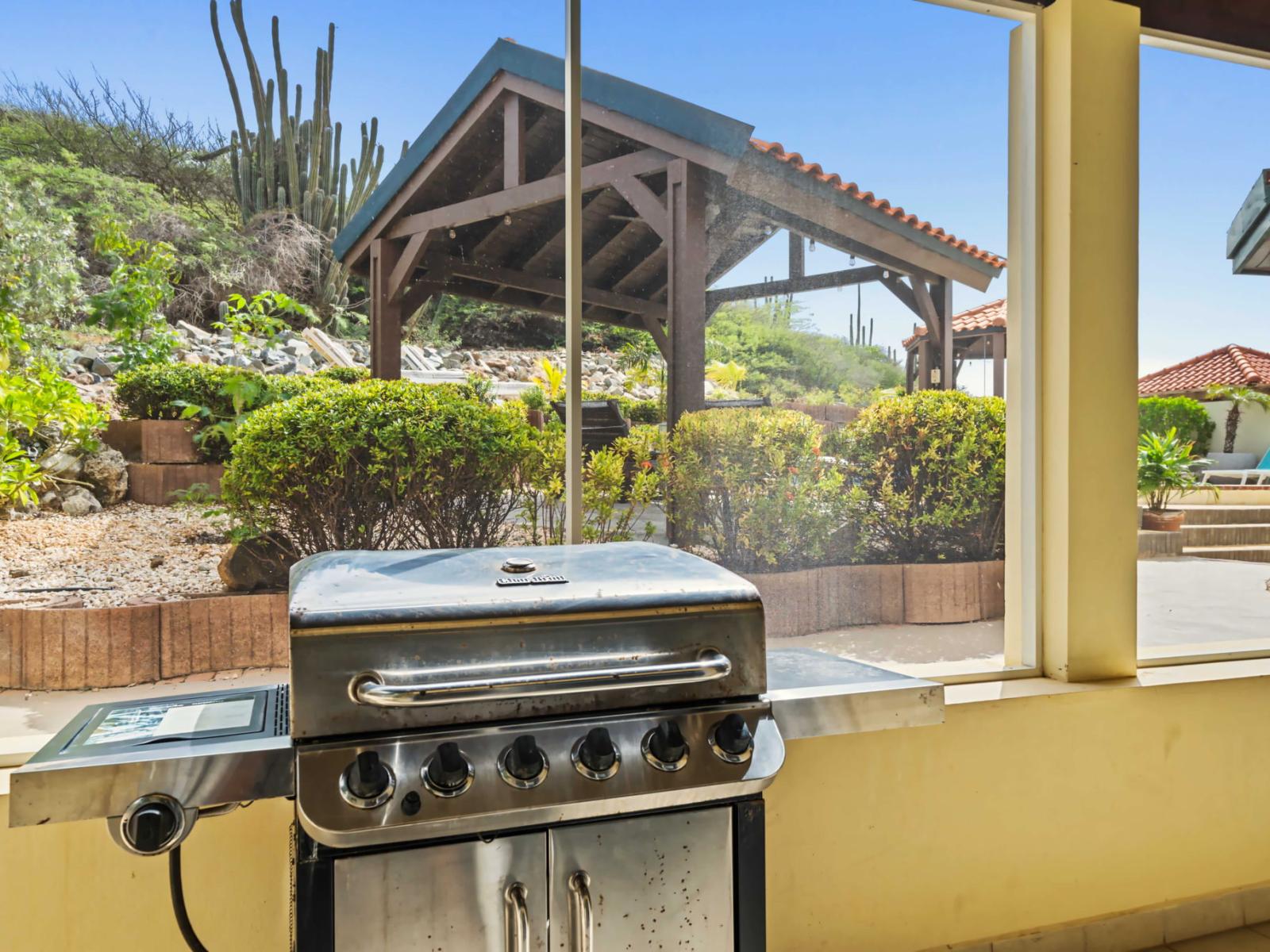 The covered patio features a BBQ grill, perfect for outdoor cooking and entertaining - The backdrop of desert plants, including tall cacti, adds a unique and picturesque element to the setting - This space is ideal for enjoying meals outdoors