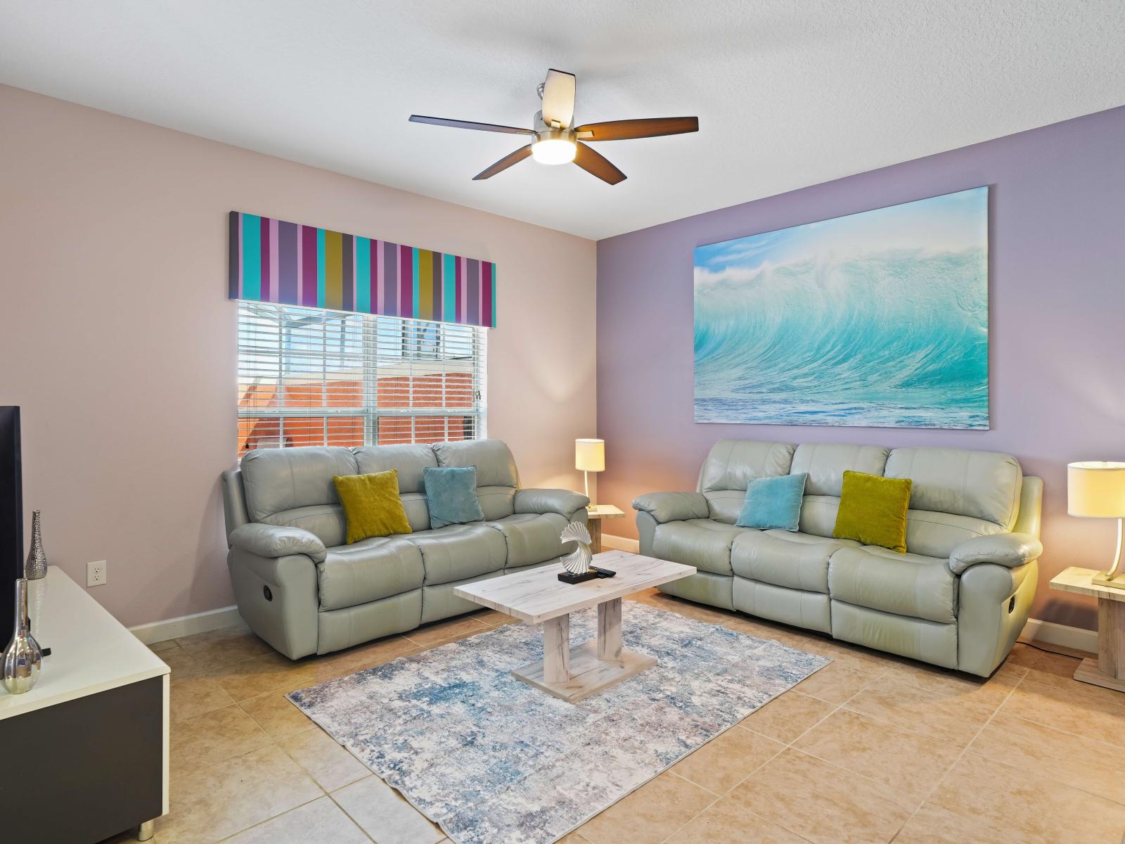 - Living room with soft gray leather sofas accented by colorful pillows and a beach-themed painting - Playful striped valance adds a vibrant touch, complementing the pastel wall colors - Soft lighting enhance the inviting and relaxed atmosphere