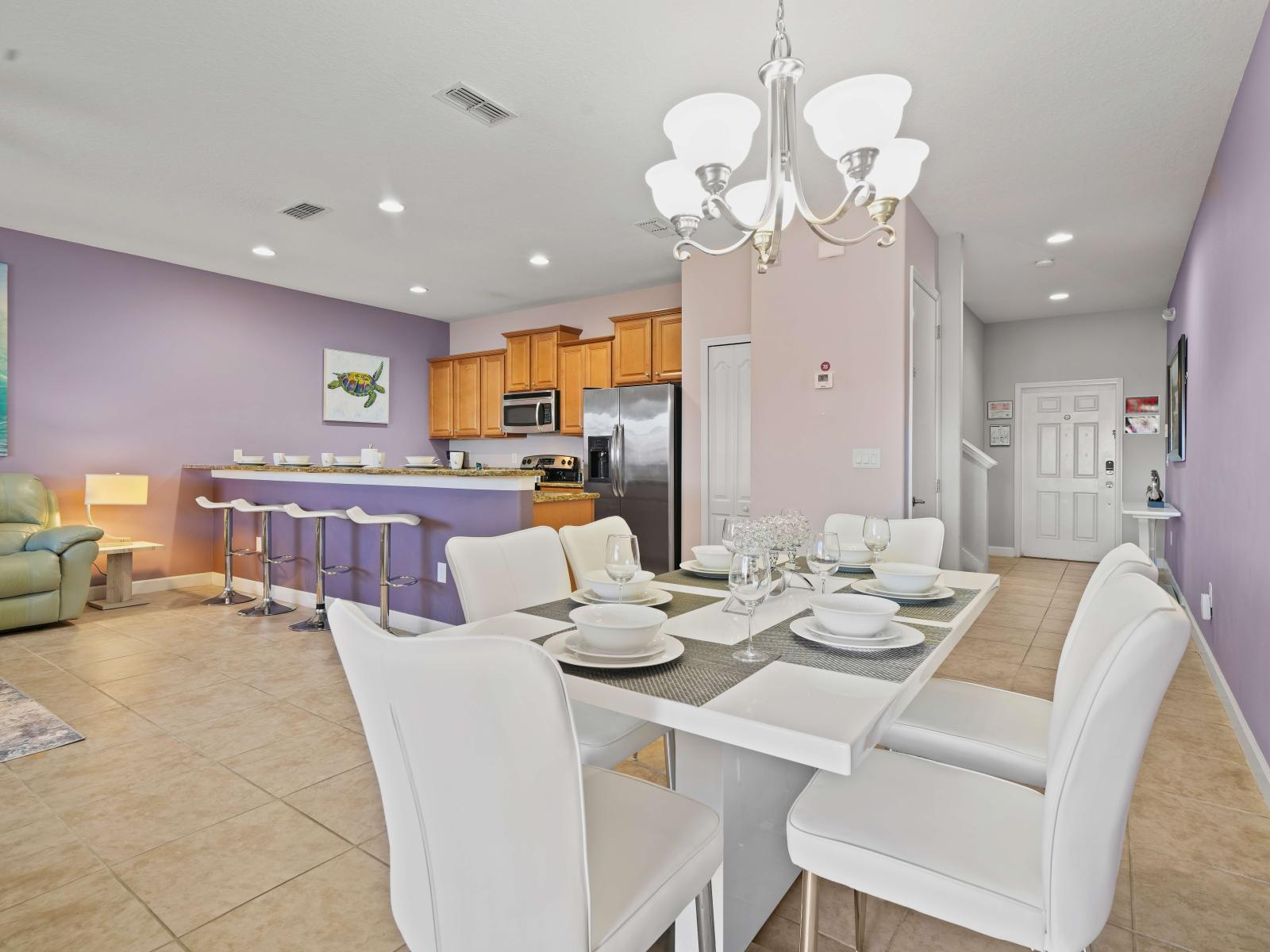 - Open-concept dining area with a modern white table and chairs, set for six - Spacious kitchen with warm wood cabinets, stainless steel appliances, and a breakfast bar with four stools - Soft purple walls add a calming and elegant touch to the space