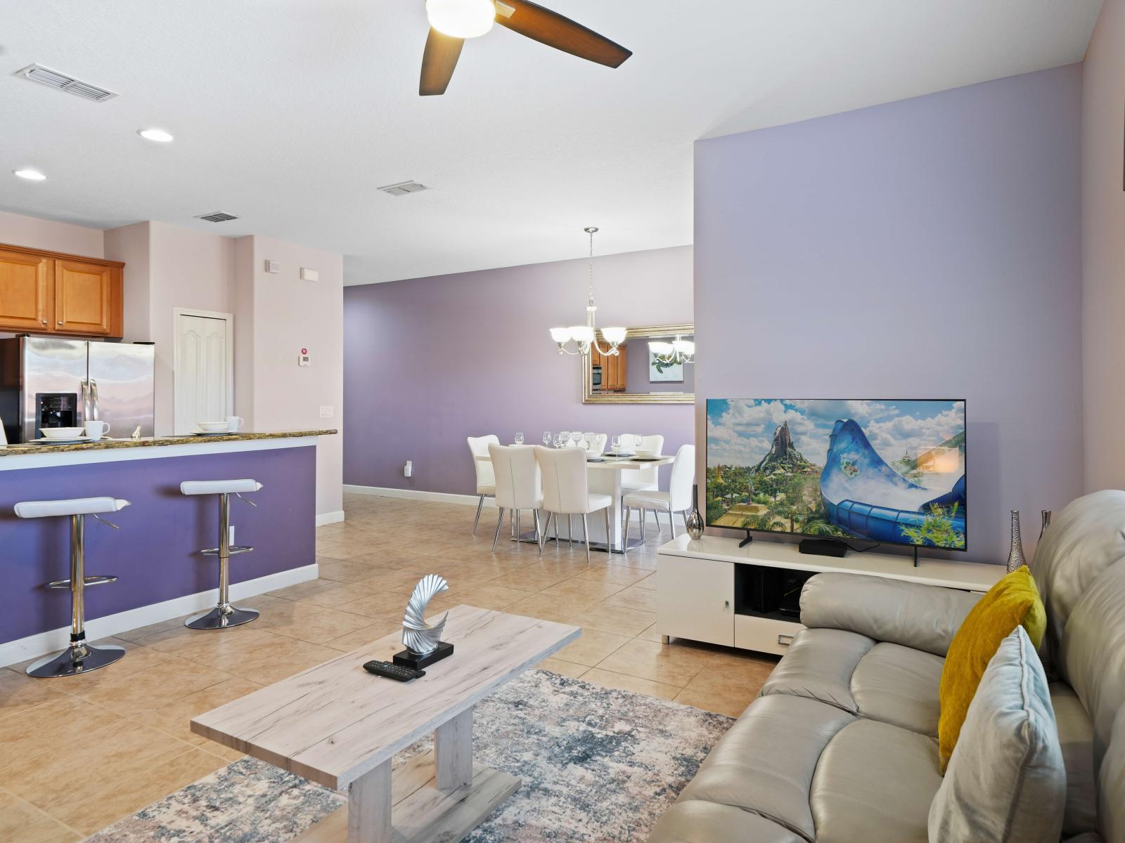 - Serene open living area with soft gray leather seating, modern decor, and a smart TV - Adjacent kitchen with a breakfast bar and stools, blending into the dining space - Light purple walls add warmth and elegance to the cohesive, inviting layout