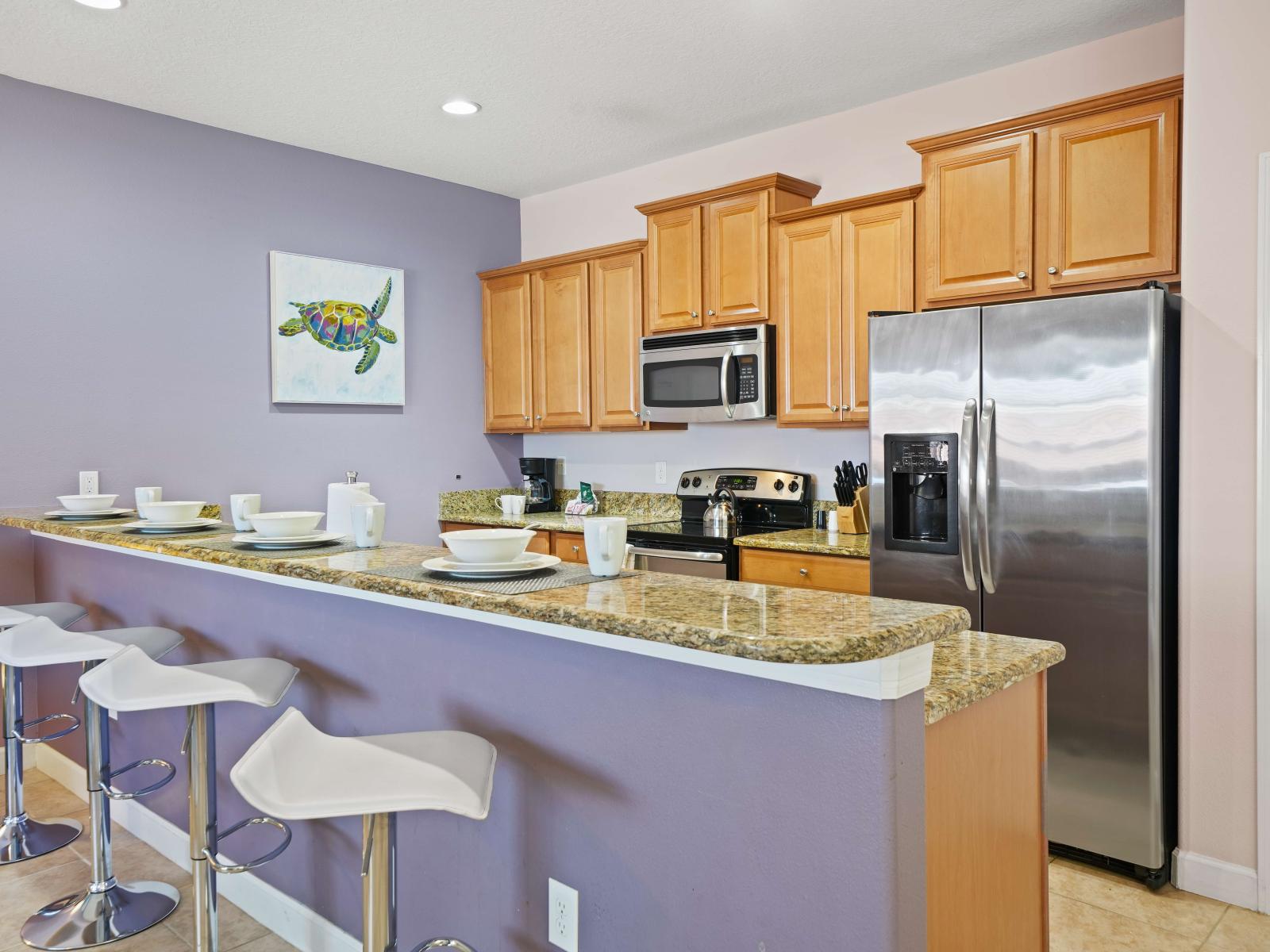 - Modern kitchen with warm wood cabinets, granite countertops, and stainless steel appliances - Breakfast bar with sleek white stools for four and a soft purple wall adds style and functionality - Artful turtle painting adds a playful, coastal touch