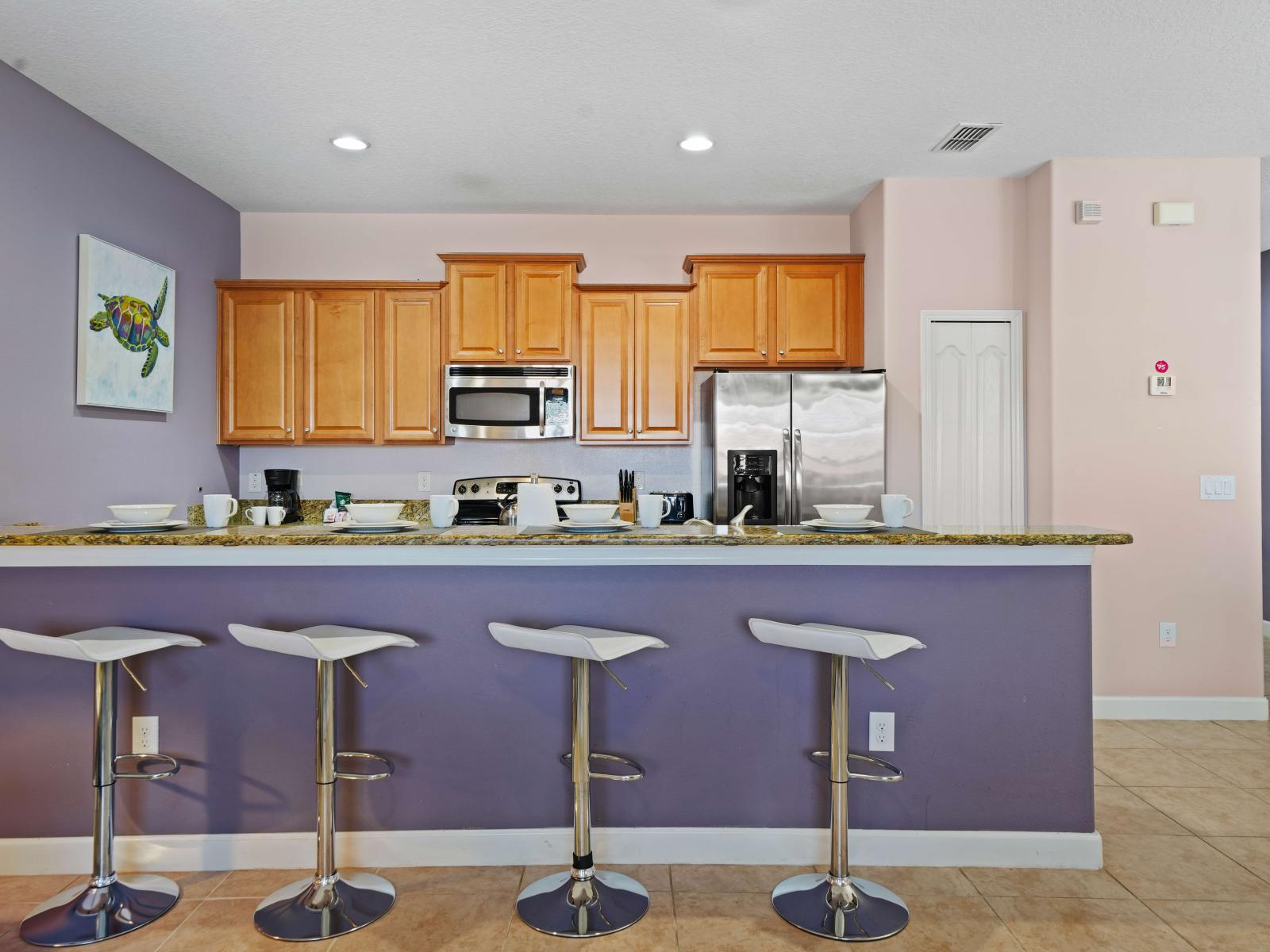 - Spacious kitchen with warm wood cabinetry, granite countertops, and stainless steel appliances - Breakfast bar with four modern white stools against a soft purple accent wall - Coastal art adds a playful touch to the stylish and functional space