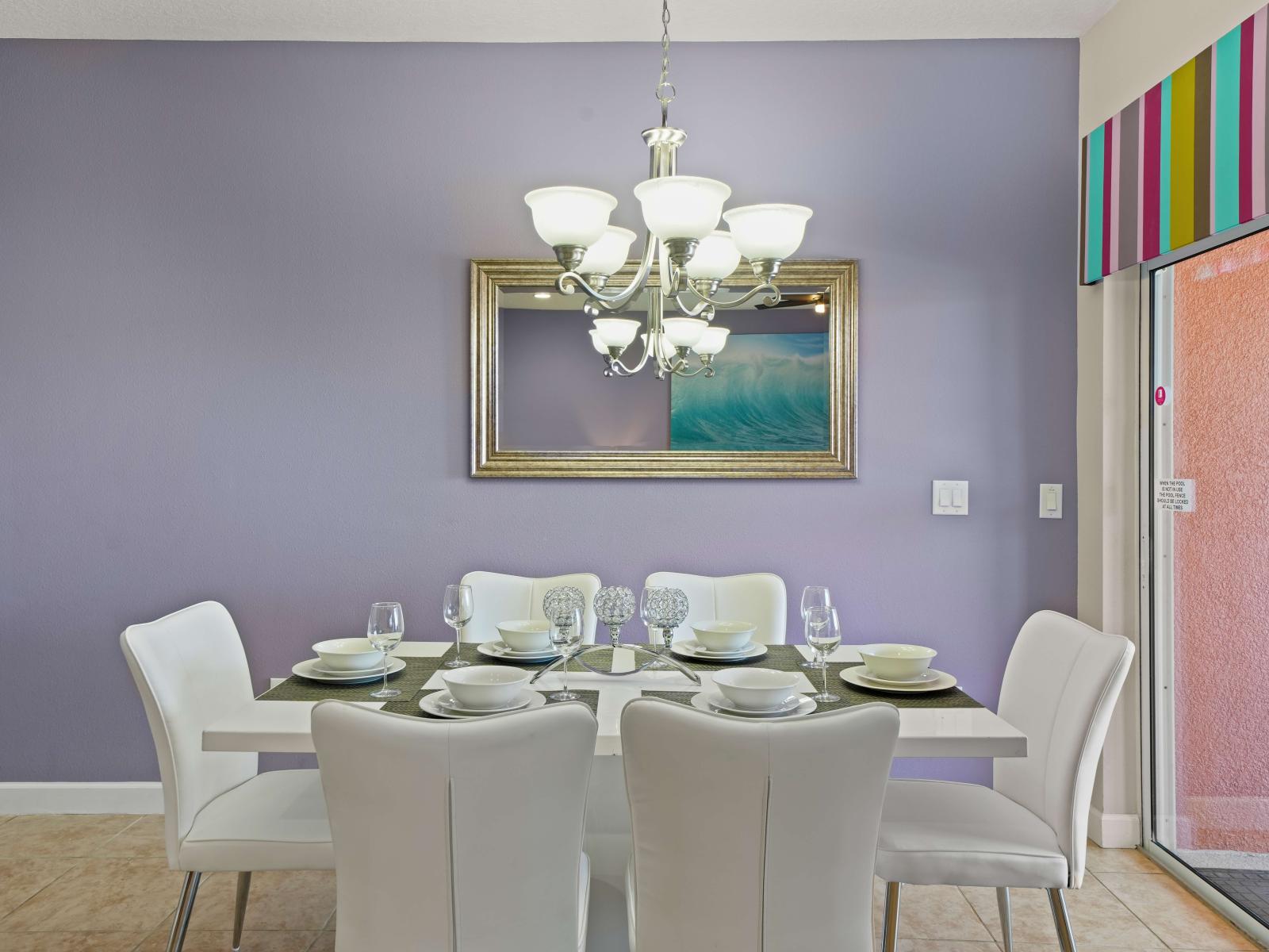 - Elegant dining area featuring a modern white table set with matching chairs and glassware - Chandelier adds a touch of sophistication, complementing the soft purple accent wall - Mirror and colorful striped valance bring a vibrant, stylish feel