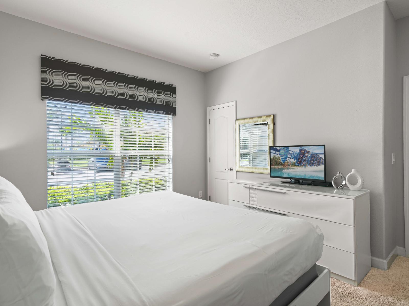 - Bright bedroom, featuring soft white linens and a sleek, minimalist design bed - Window with a stylish striped valance lets in natural light, brightening the space - Dresser with a flat-screen TV and modern decor creates a cozy, functional retreat