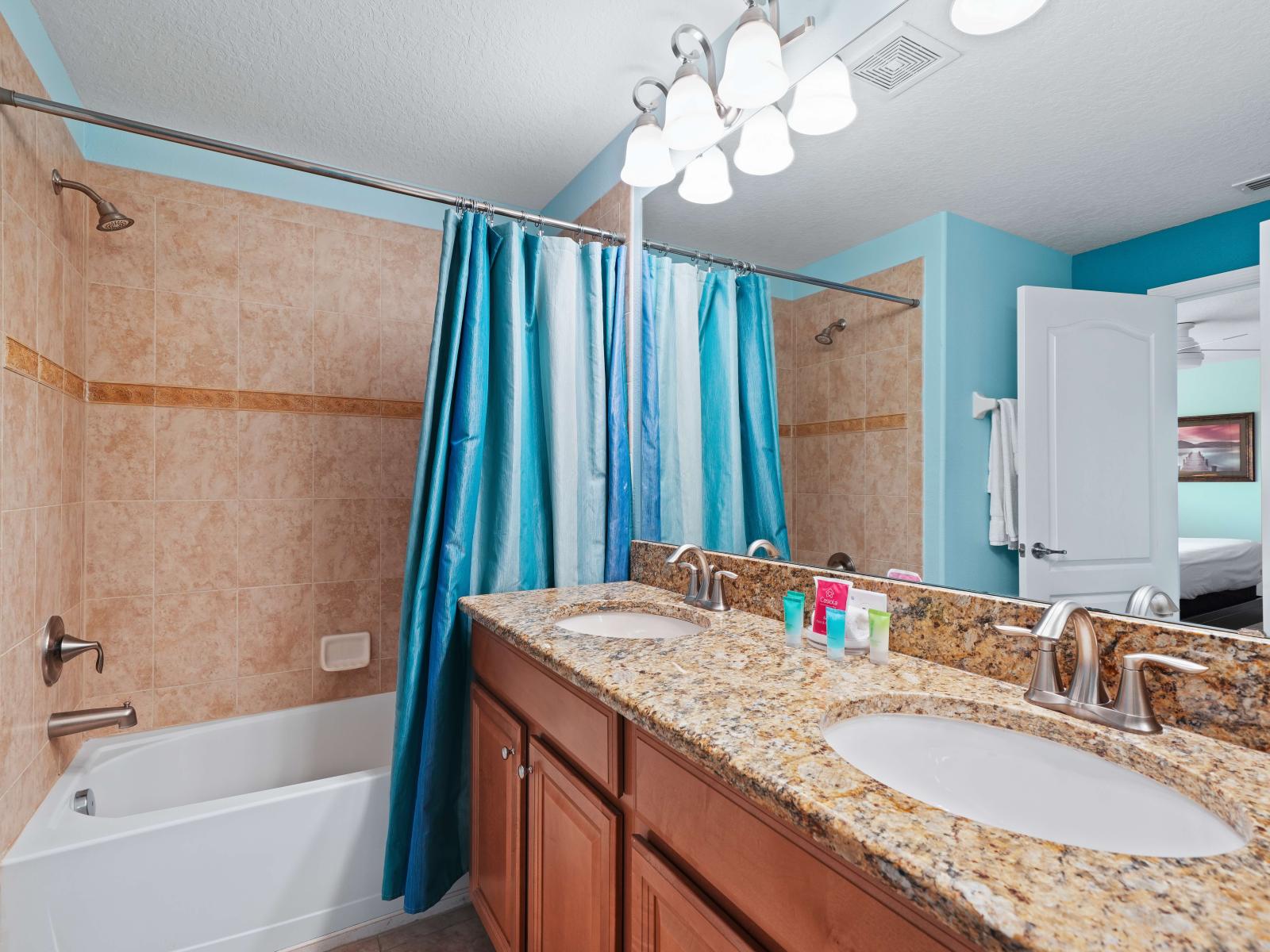 - Bright bathroom featuring a double sink with granite countertops and a matching wood vanity - Tiled bathtub and shower combination with a blue curtain adds a splash of color - Elegant lighting and a spacious layout create a fresh, functional space